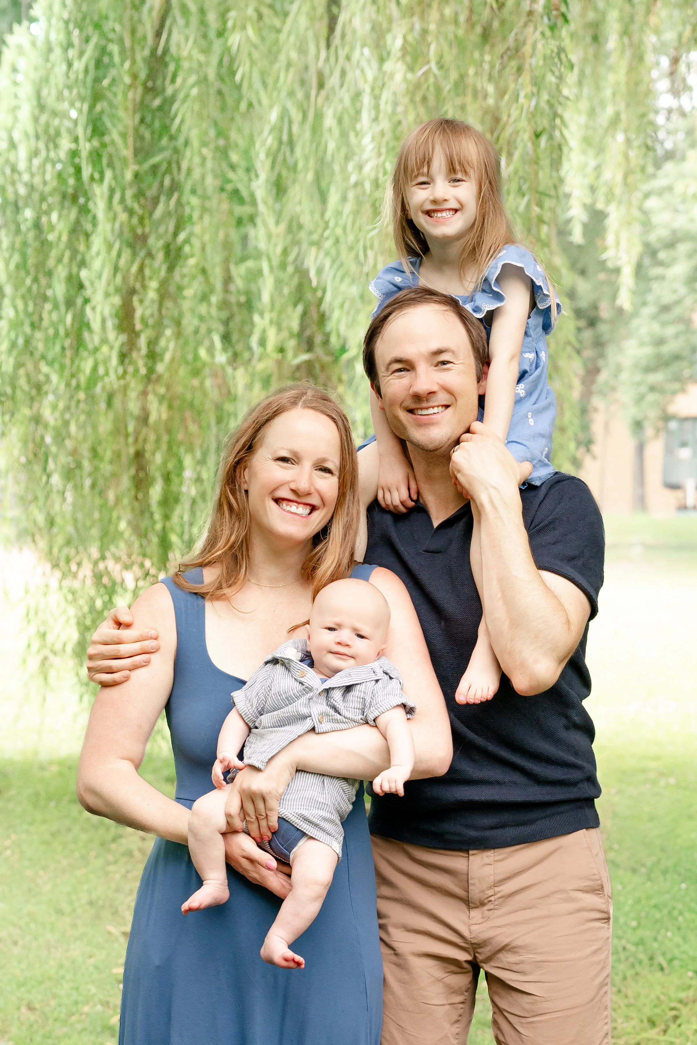 A happy family of four outdoors in a park with green grass and trees, including a woman, a man, a young girl on his shoulders, and a baby in the woman's arms. Located in Glenview IL.
