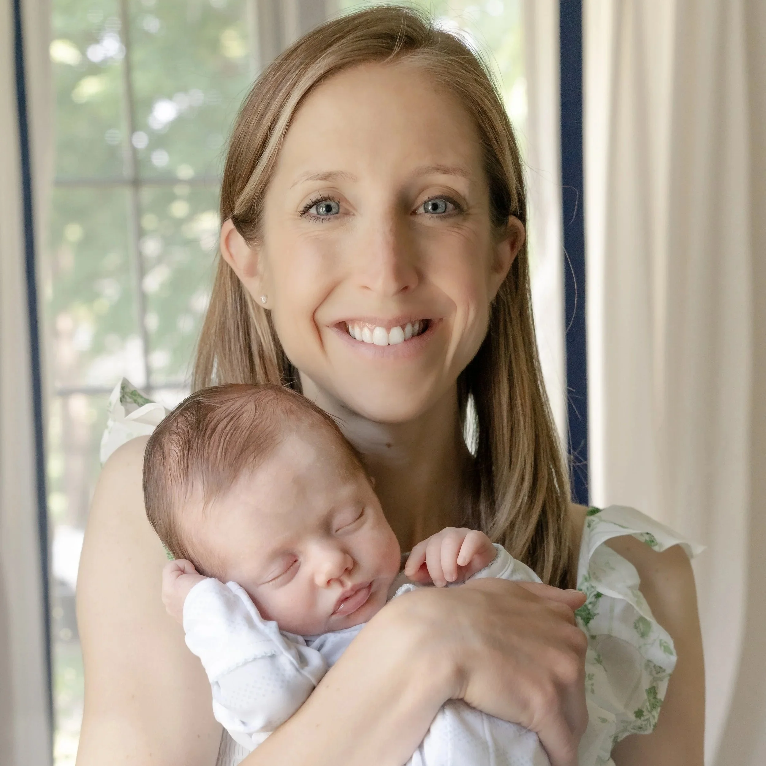 A woman with light brown hair and blue eyes holding a sleeping baby with reddish-brown hair, both smiling, near a window with sheer curtains and trees outside located in Winnetka IL.