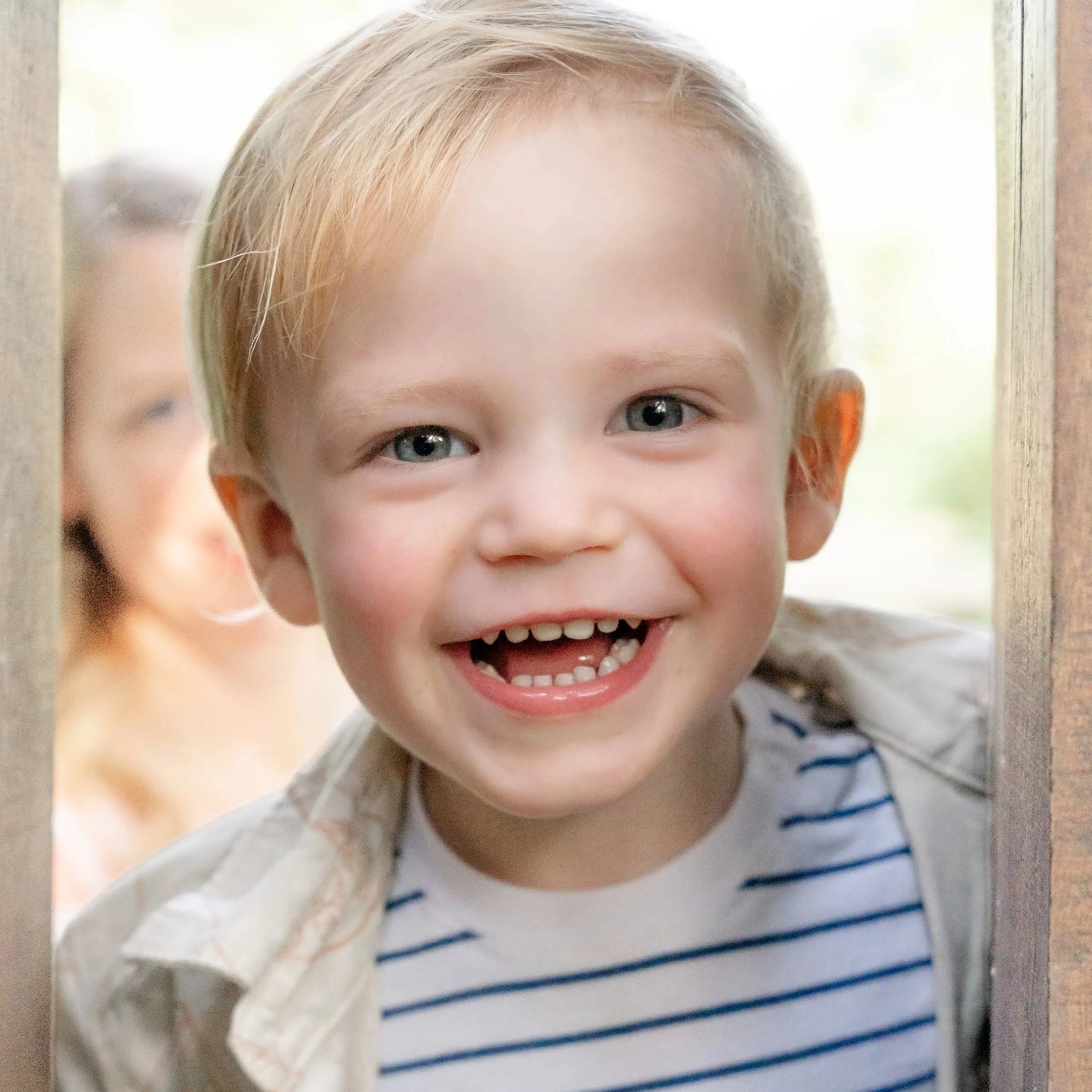 Close-up of a young boy with blond hair and blue eyes, smiling, wearing a striped shirt, partly hidden behind a wooden door located in Glenview IL.