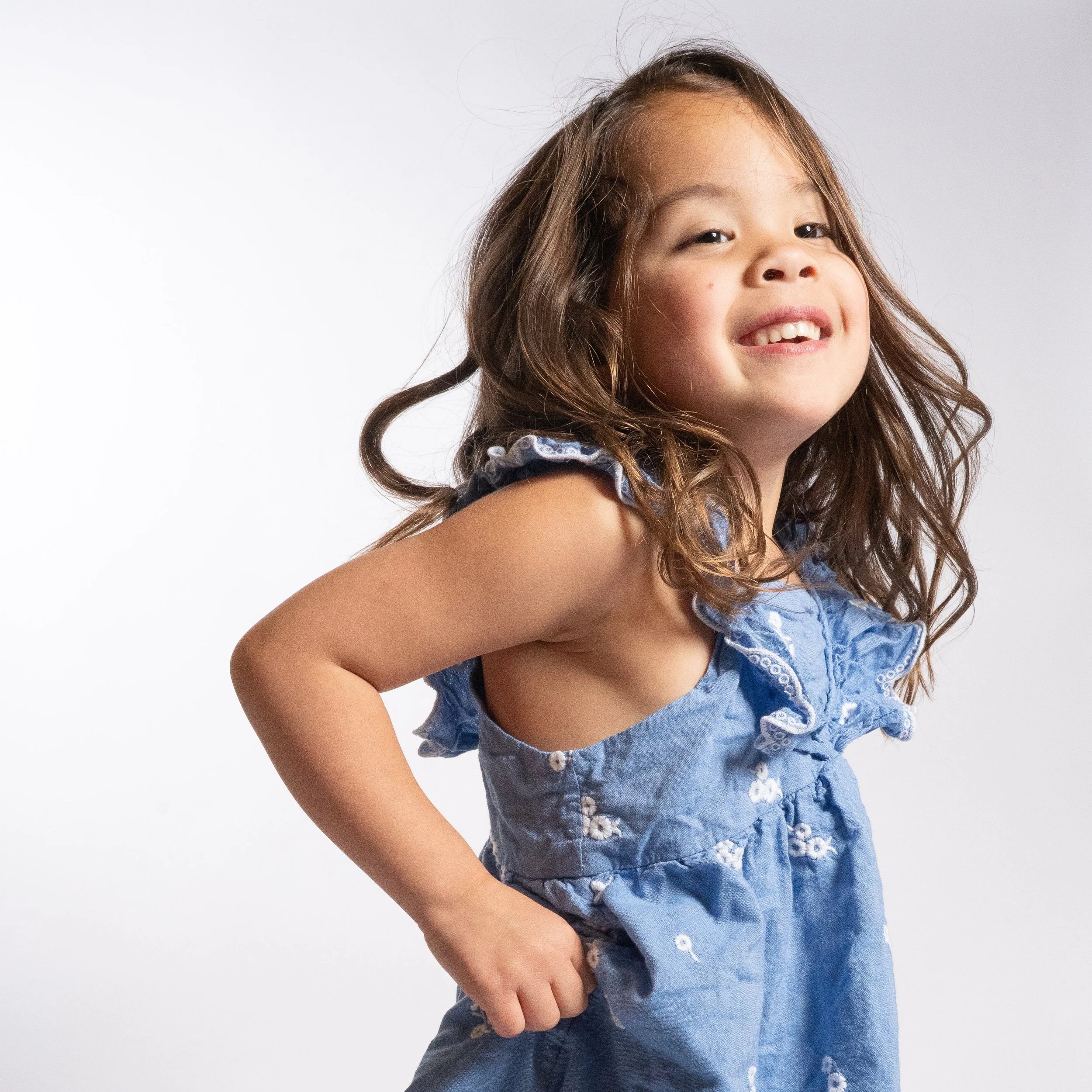 Close-up of a young girl with curly hair smiling and showing her teeth, wearing a blue dress with white floral embroidery located in Glenview IL.
