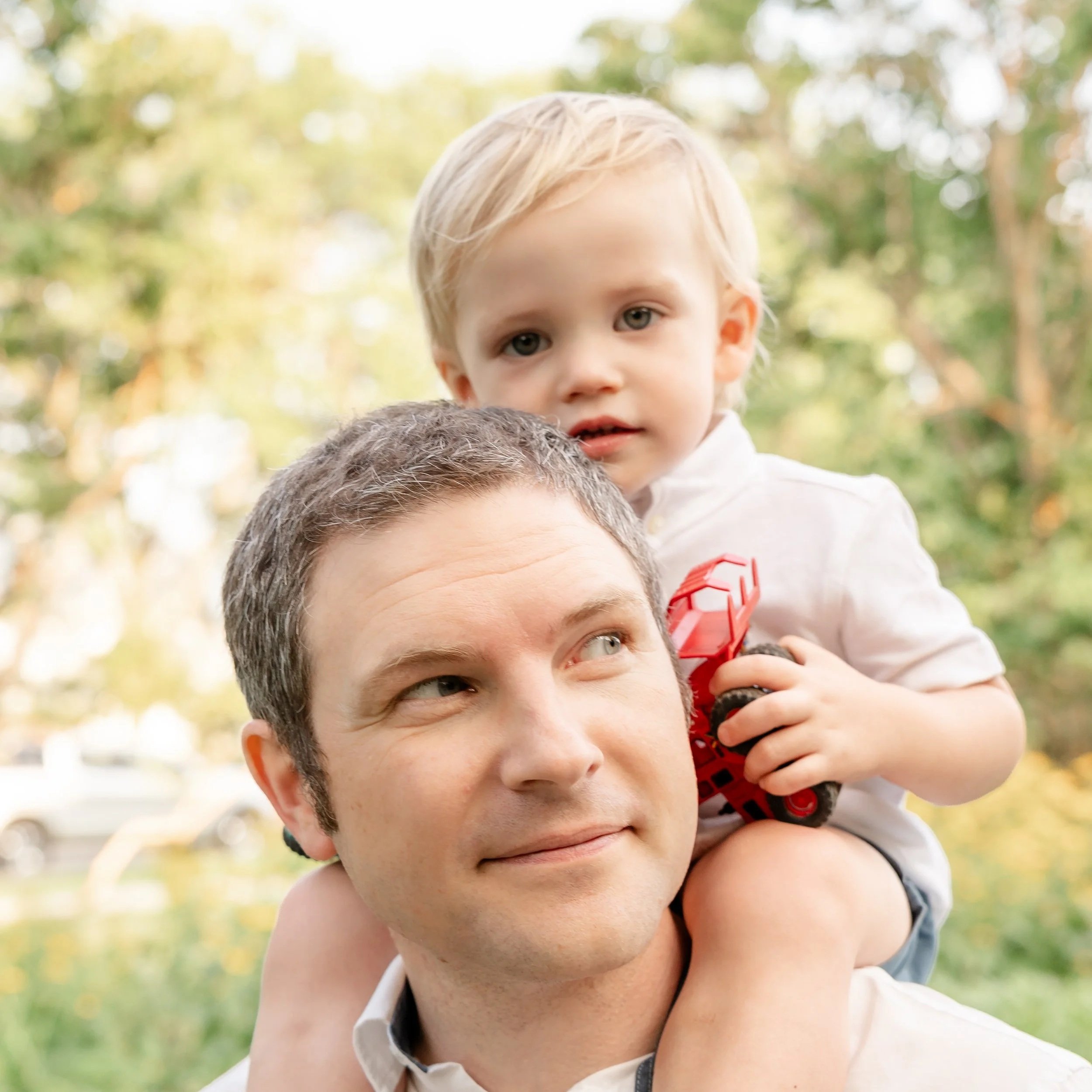 A young boy with blonde hair and blue eyes is sitting on a man's shoulders outdoors with blurred trees and sunlight in the background located in Glenview IL.