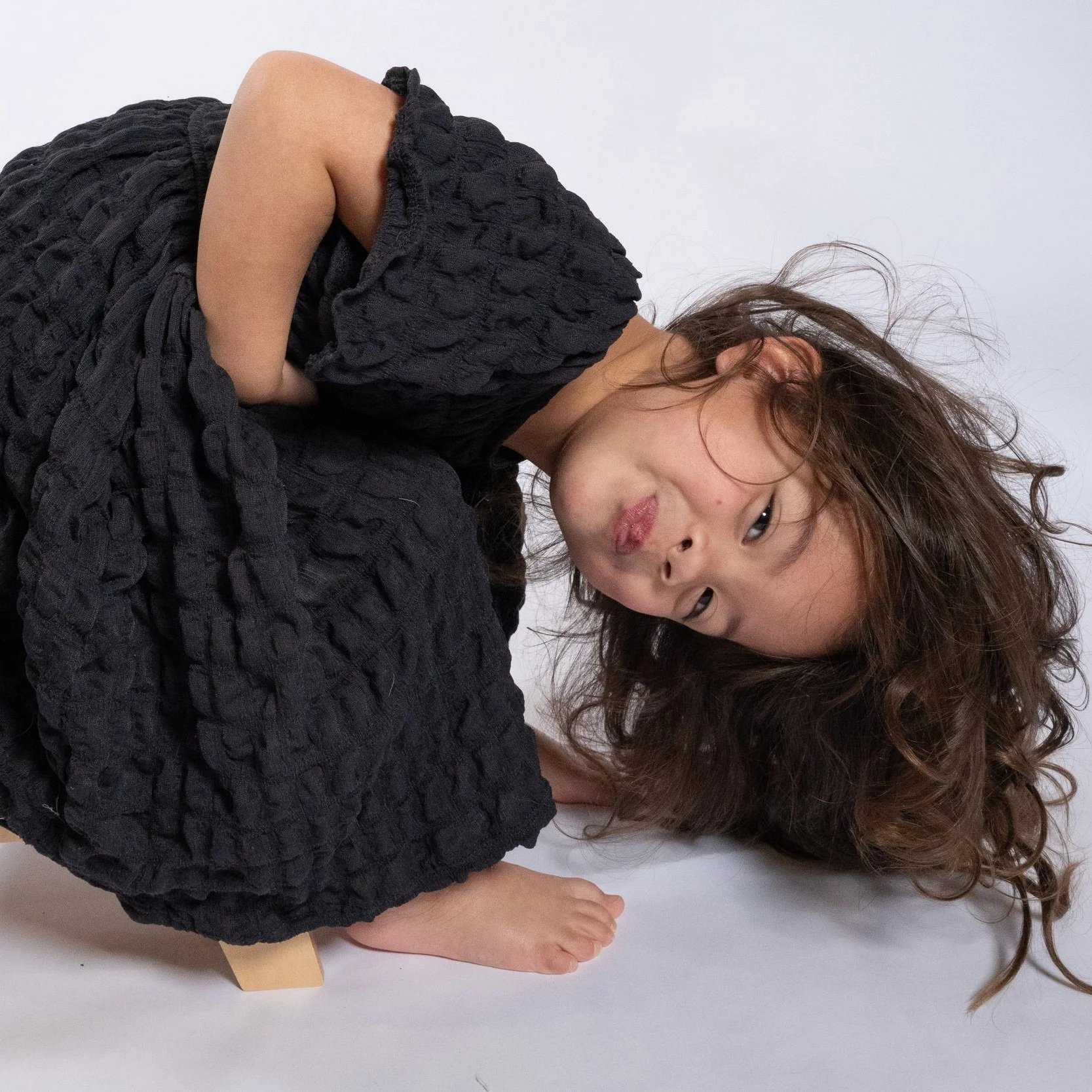 A woman with long, wavy hair wearing a black textured dress, lying on the floor with her head tilted to the side and holding a foot with her hand located in Glenview IL.