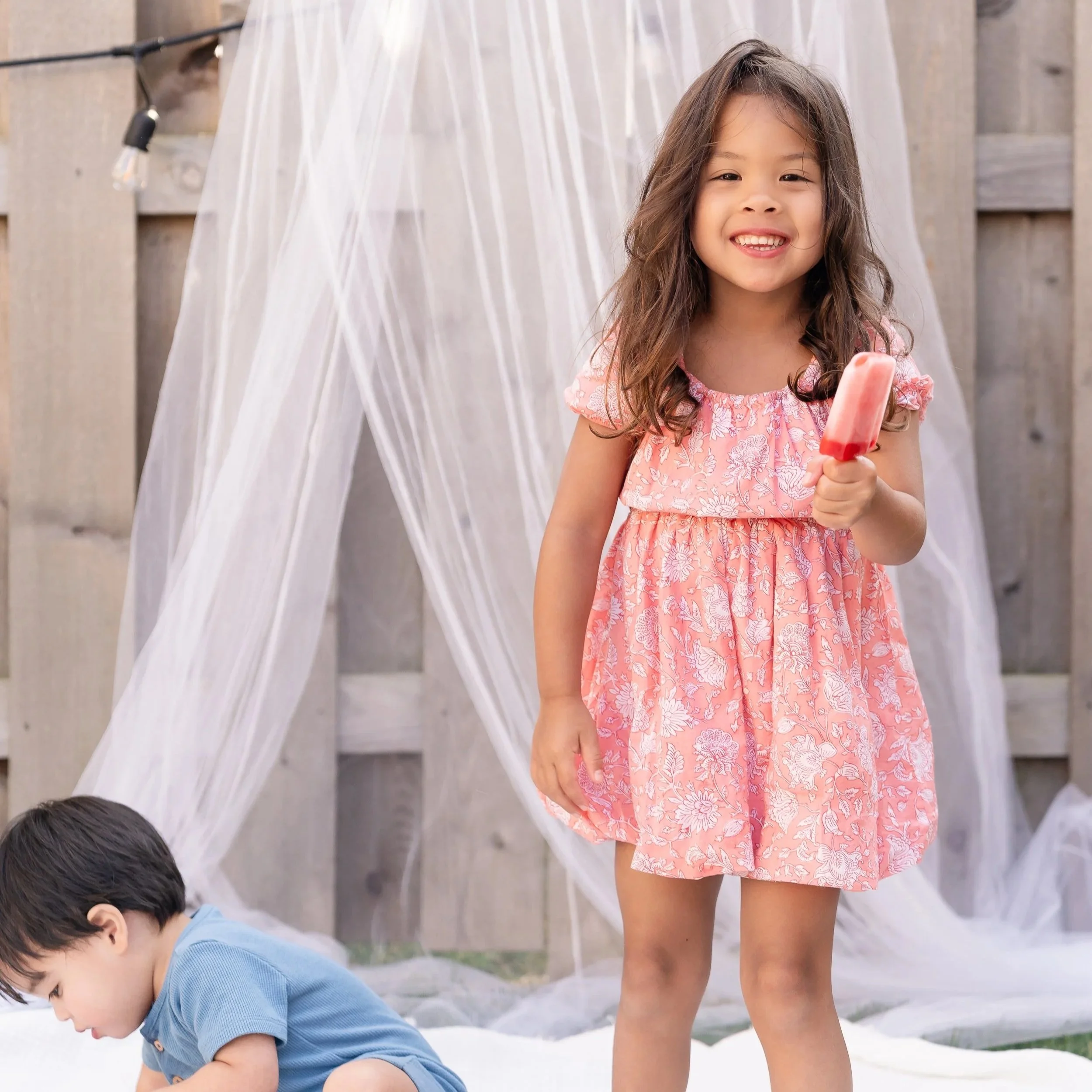 A young girl in a pink floral dress holding a red popsicle, smiling, standing outdoors in front of a white sheer curtain backdrop, with a boy in a blue shirt partially visible and crawling on the ground located in Glenview IL.