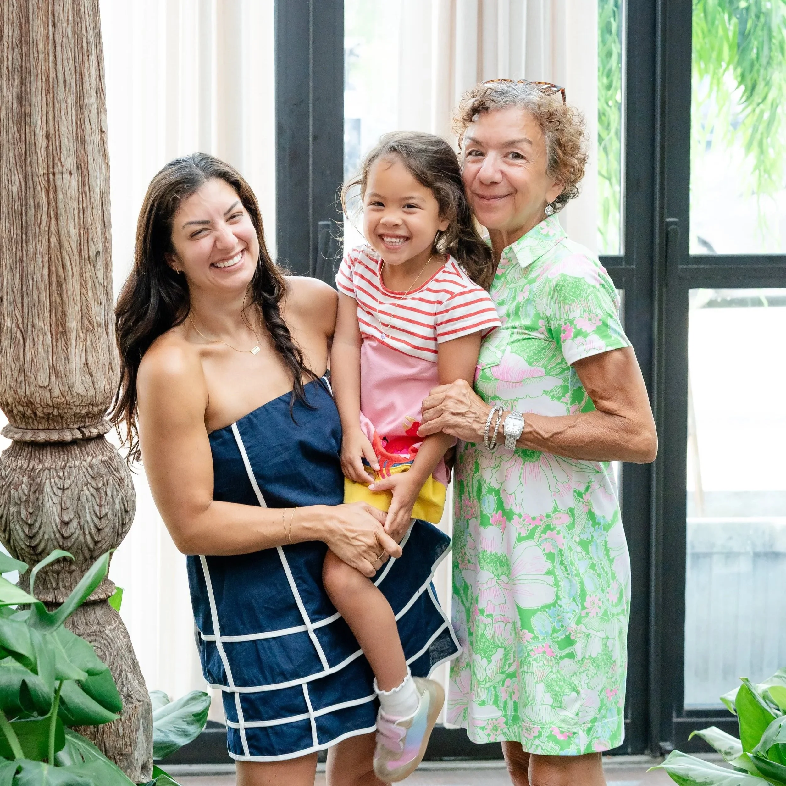 Three generations of women, a young woman, an older woman, and a young girl, smiling and standing close together indoors near large windows and a tree, with green plants visible located in Glenview IL.