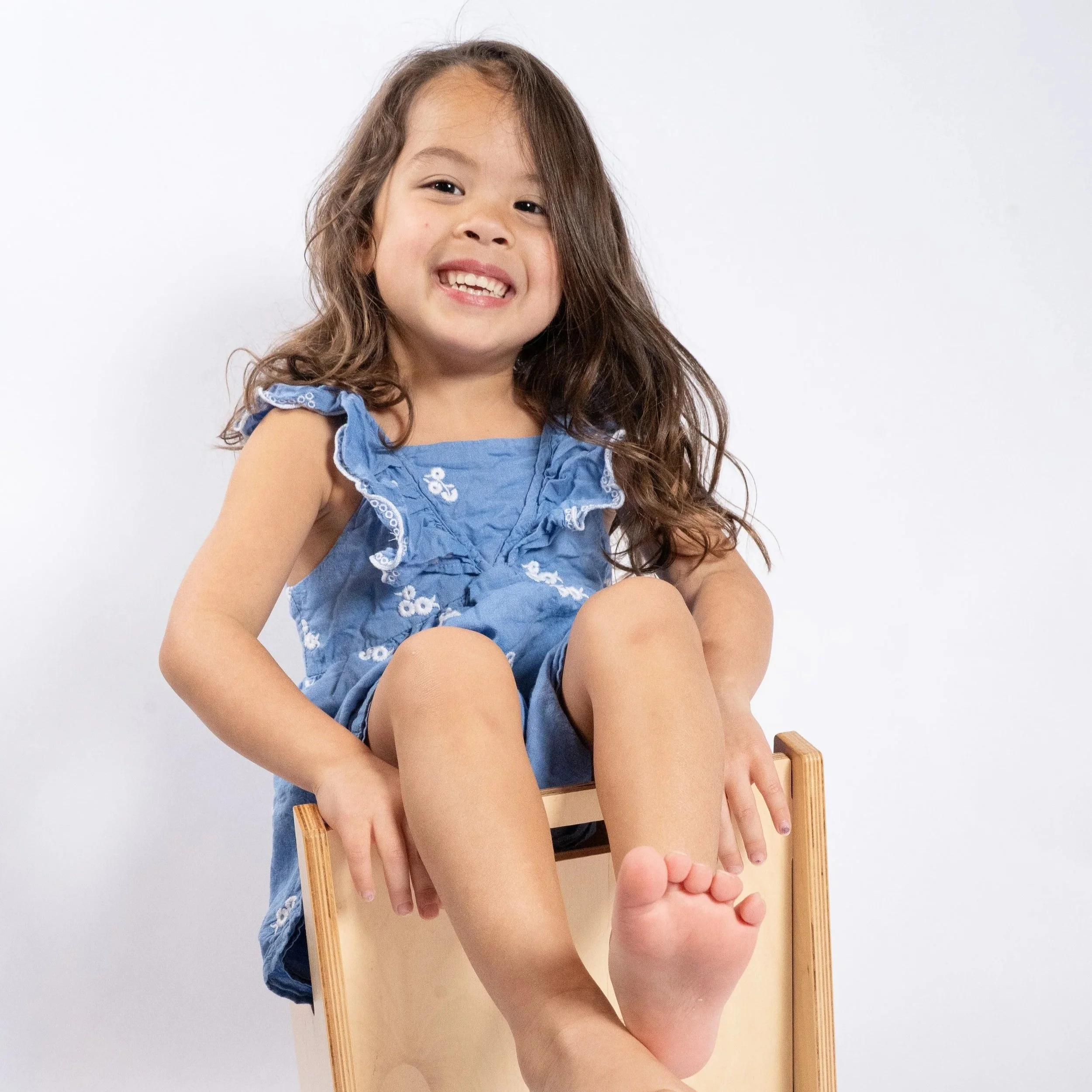 Young girl with long curly hair sitting on a wooden chair, smiling and showing her teeth, wearing a blue sleeveless dress with white floral embroidery, against a plain white background in Glenview IL.