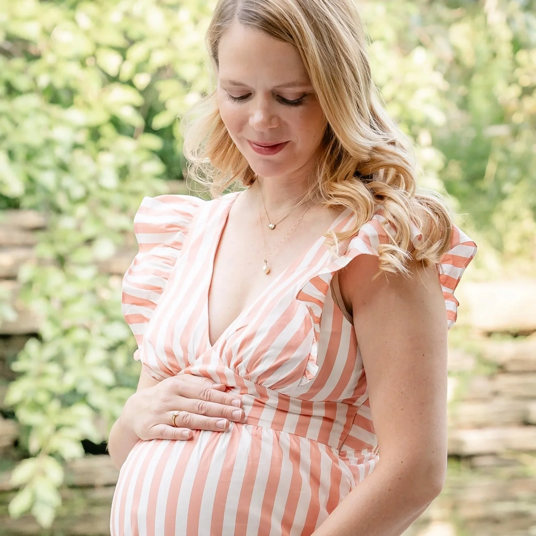 A woman with blonde hair wearing a pink and white striped dress, looking down outdoors during daytime, with greenery in the background in Glenview IL.