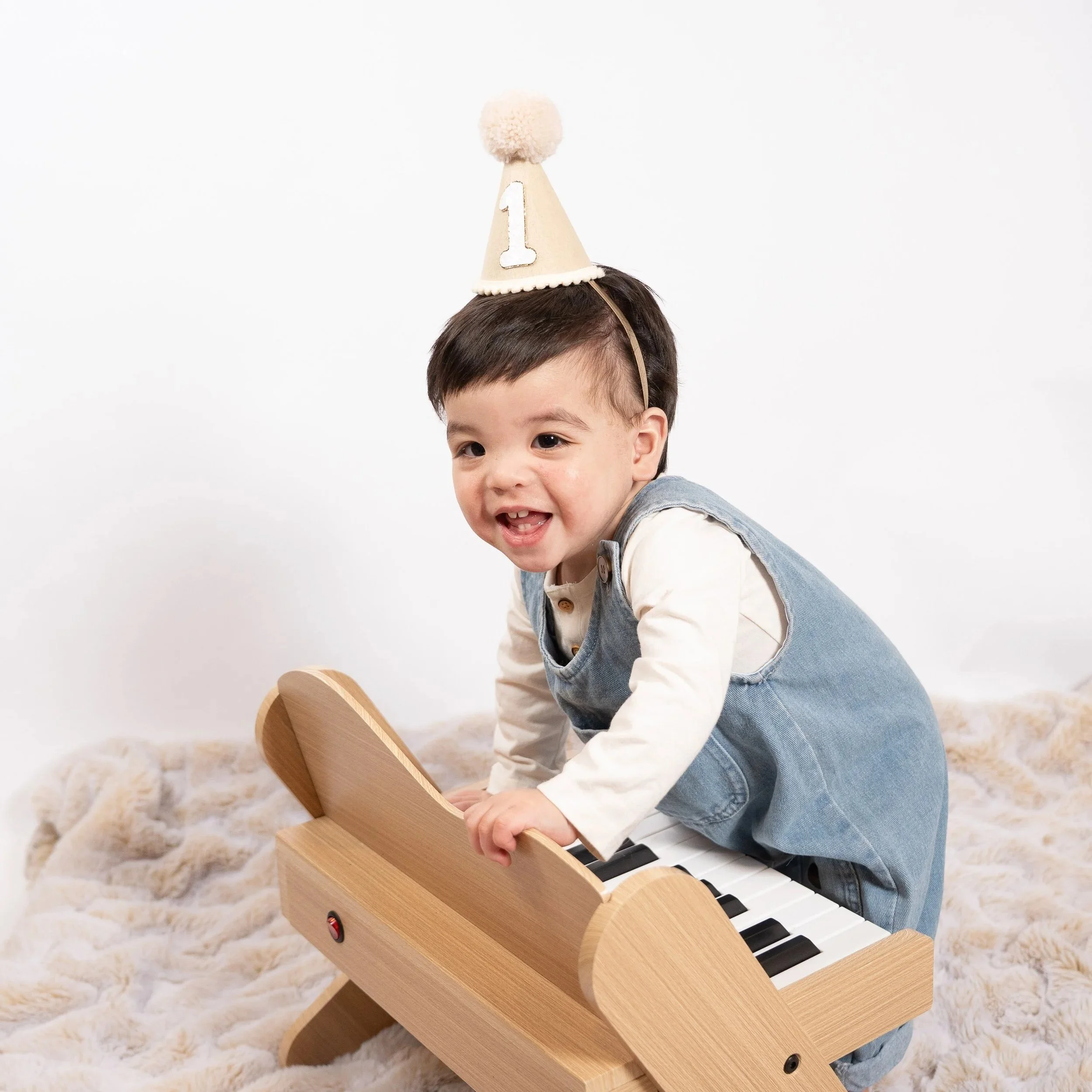 Young boy wearing a birthday hat with the number 1 on it, playing a wooden toy piano on a fluffy rug in Glenview IL.