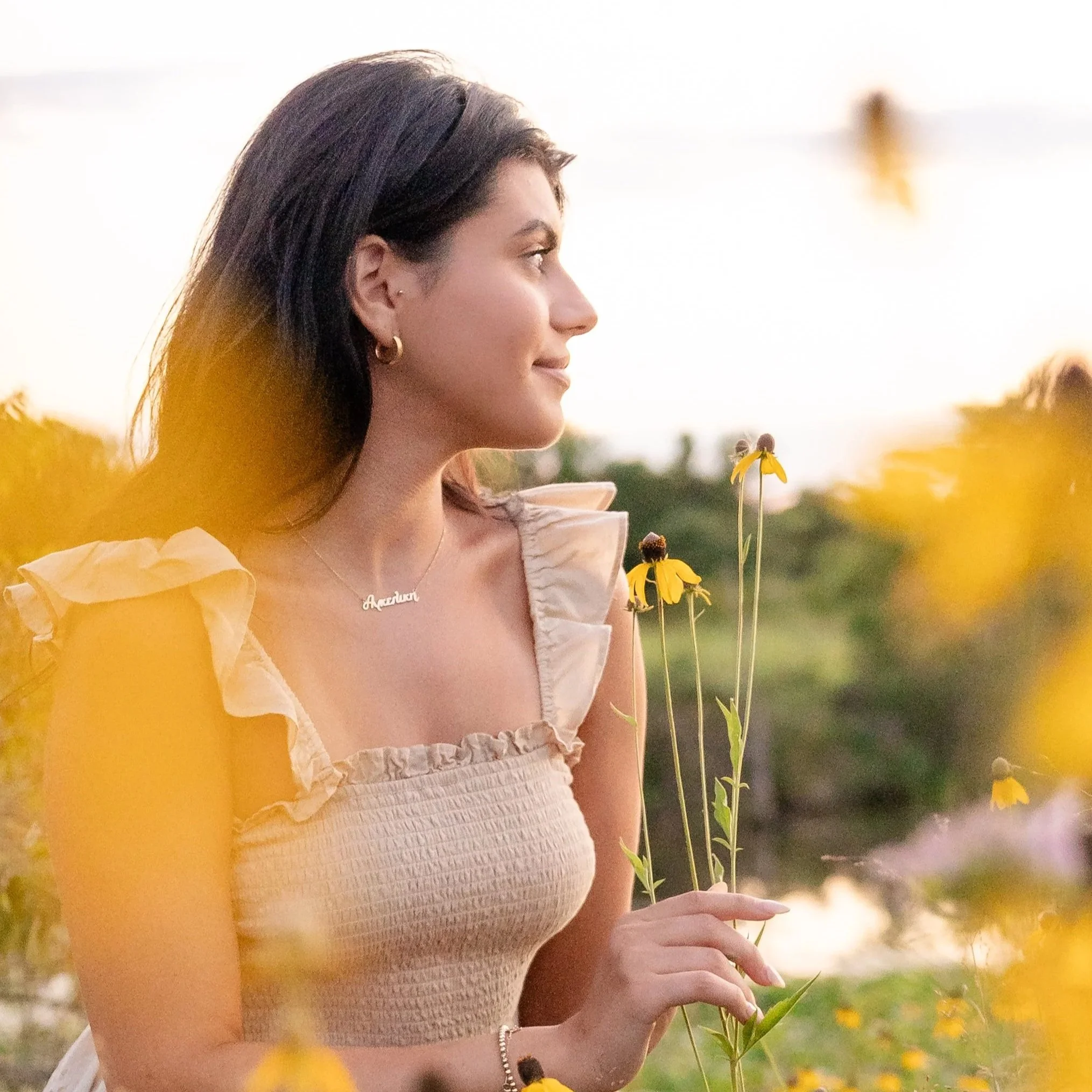 A woman in a white top and ruffled sleeves stands outdoors holding a flower and surrounded by yellow flowers, with a blurred background of trees and sky in Glenview IL.