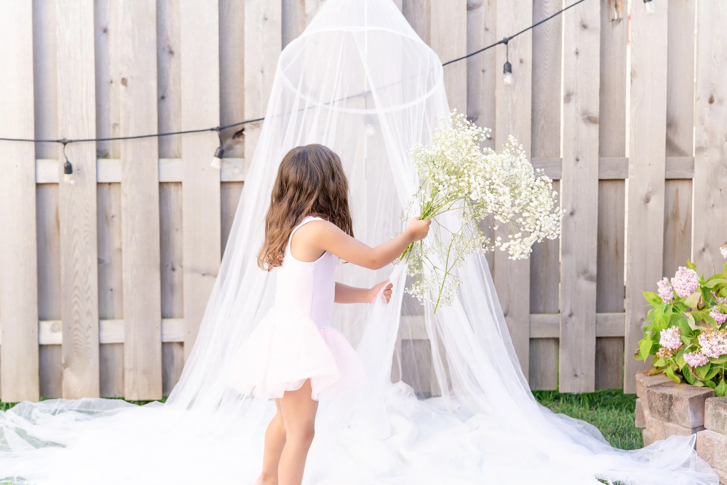 Child in a white dress holding a bouquet of white flowers near a wooden fence, with a canopy and string lights in the background in Glenview IL.