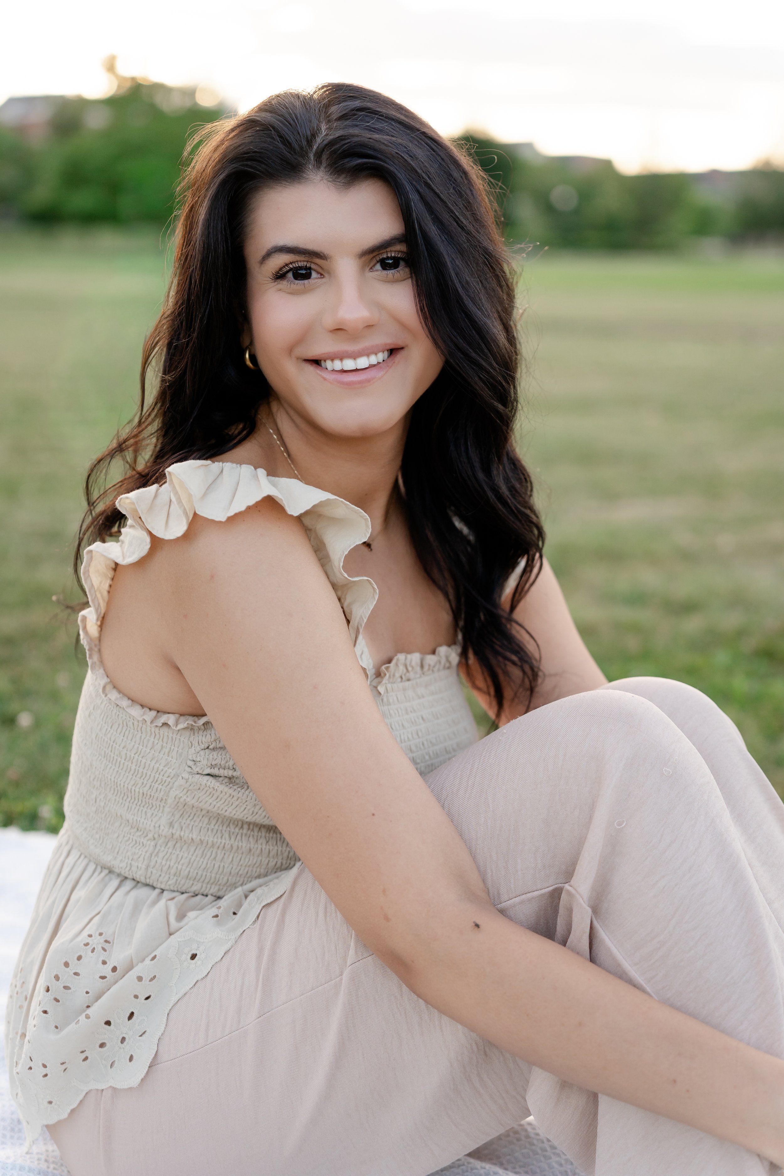 Young woman with dark wavy hair, smiling, sitting outdoors in a grassy area with trees in the background, wearing a cream-colored ruffled sleeveless top and light-colored pants in Glenview IL.
