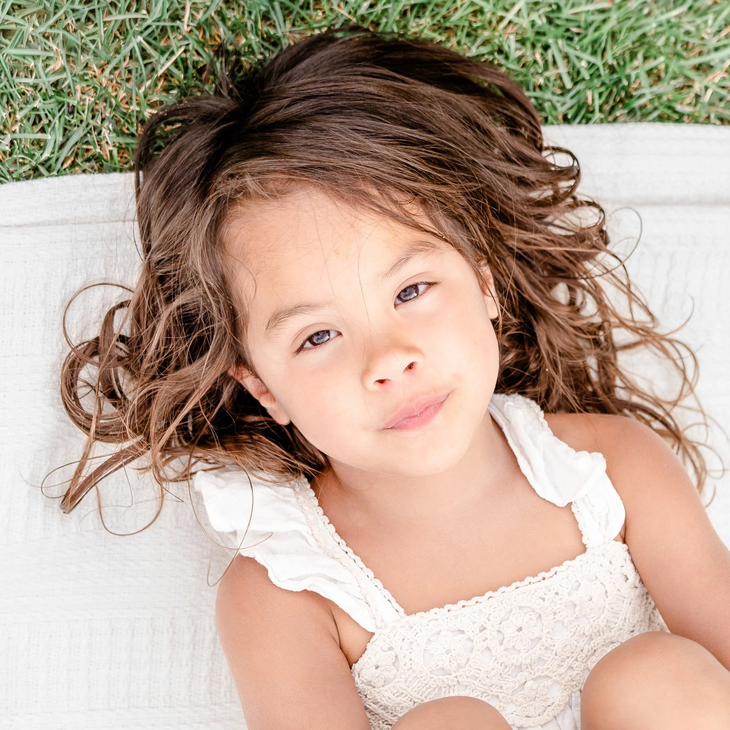 A young girl with curly brown hair and brown eyes lying on a white surface outdoors with grass in the background located in La Grange and Hinsdale, IL.