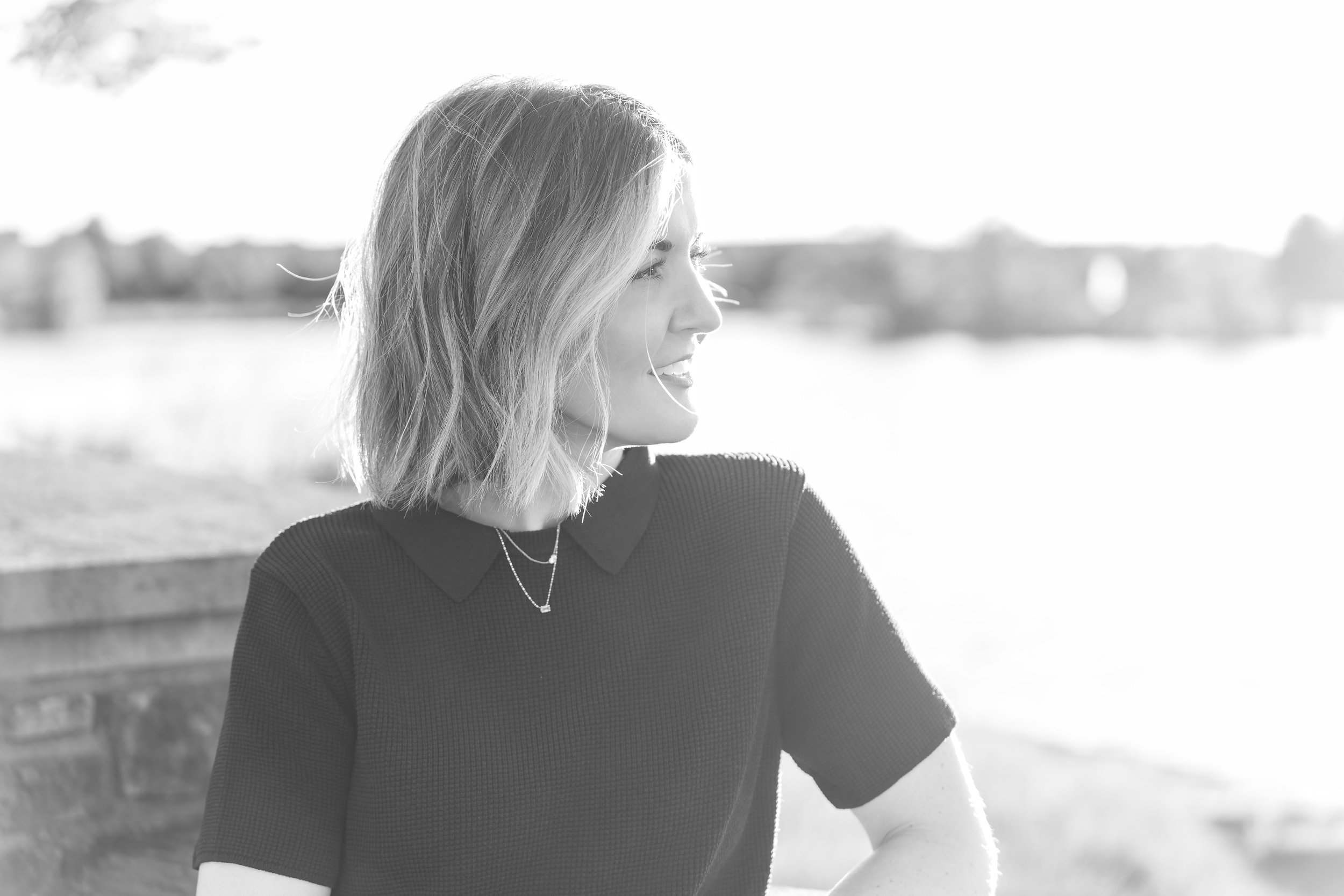 A woman with shoulder-length hair wearing a black top and a necklace, smiling and looking to the right, outdoors with a blurred background and sunlight.