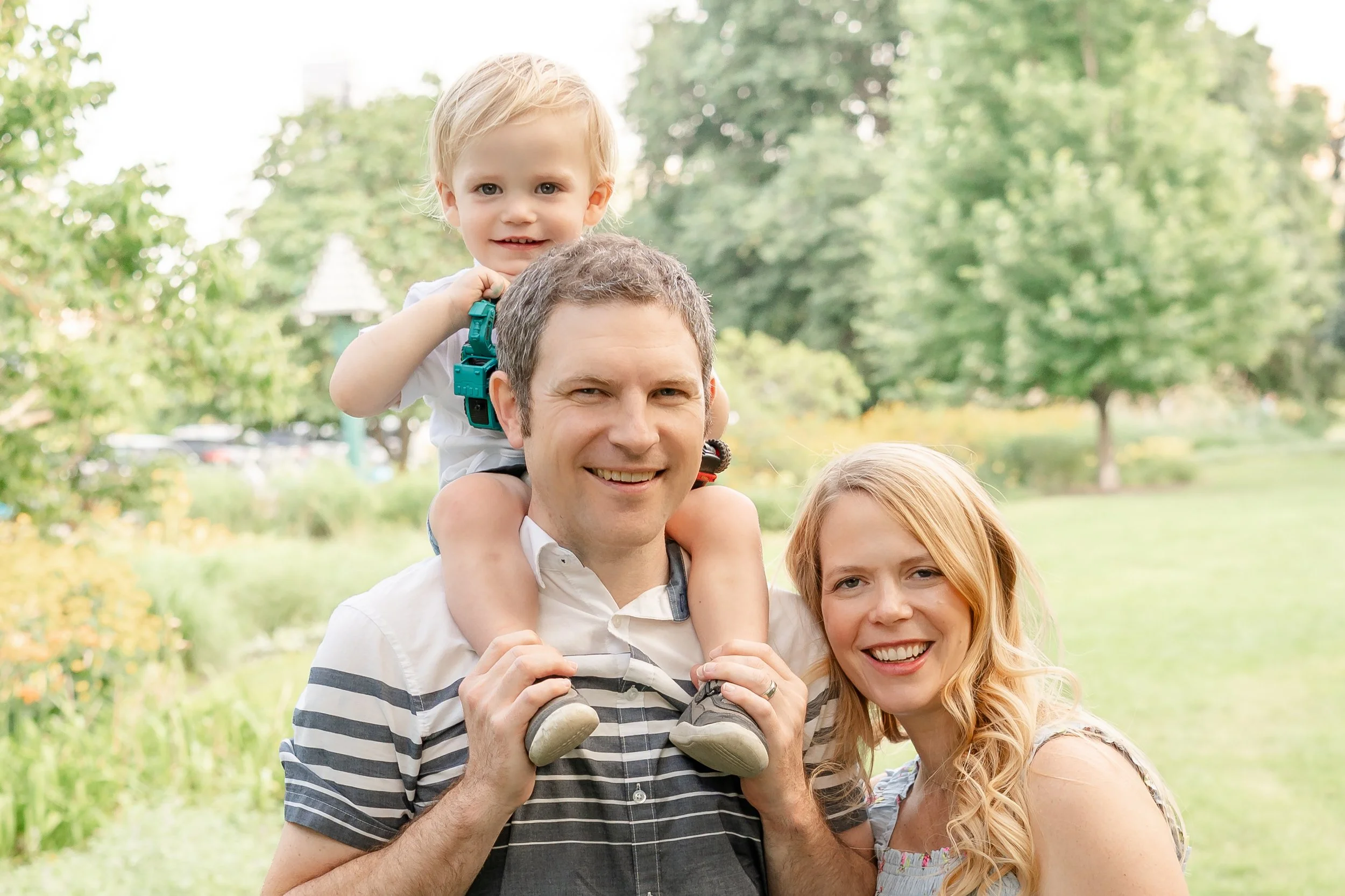 A man with short gray hair carrying a young boy on his shoulders, outdoors in a park with green trees in the background. The boy has blonde hair and is smiling, holding onto the man’s head located in Lincoln Park Chicago.