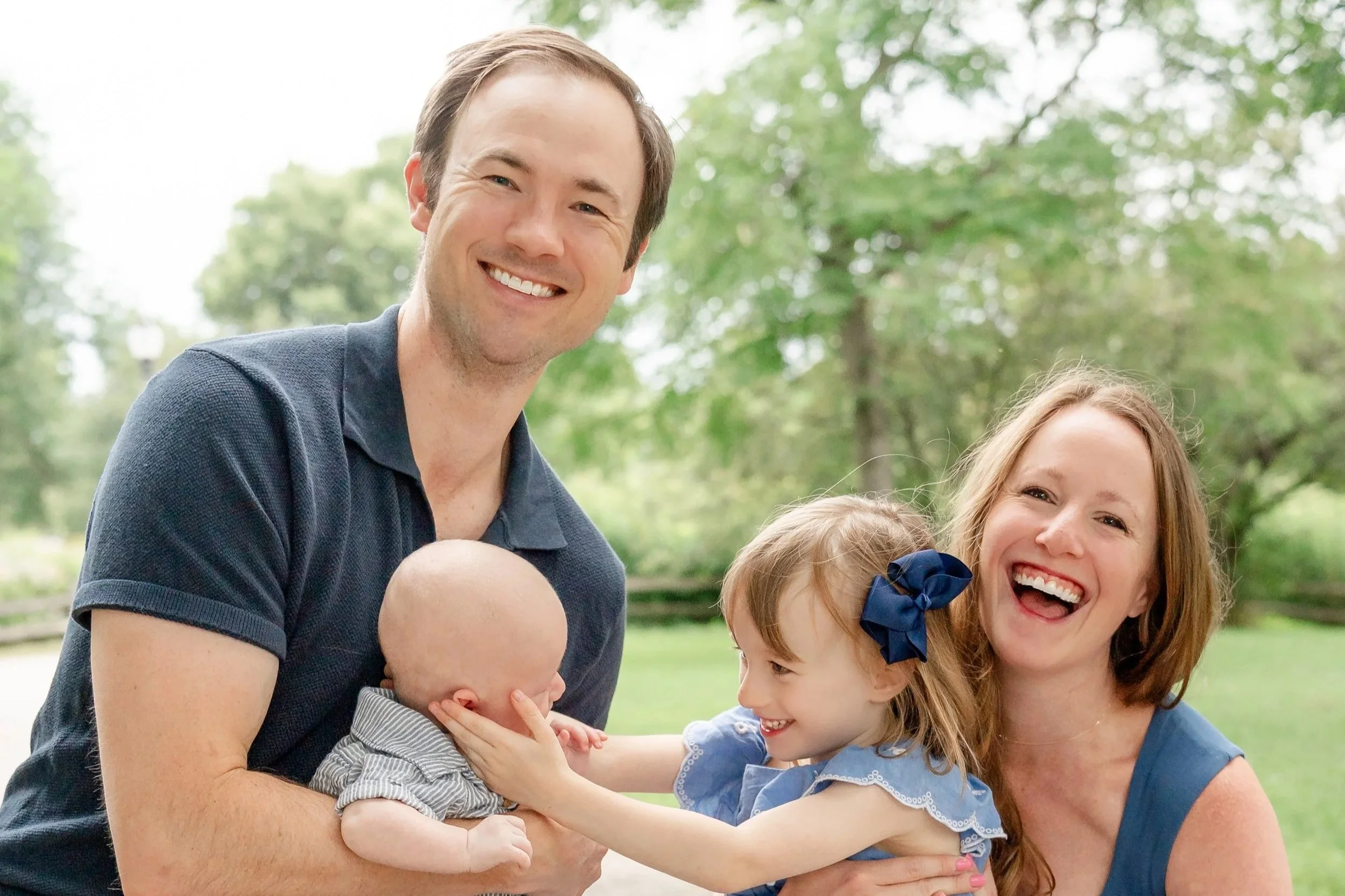 Smiling man with short brown hair wearing a black polo shirt, hugging a baby, and smiling woman with long red hair and a blue dress, with two young girls smiling and one girl wearing a blue bow headband, outdoors in chicago Glenview at a park.