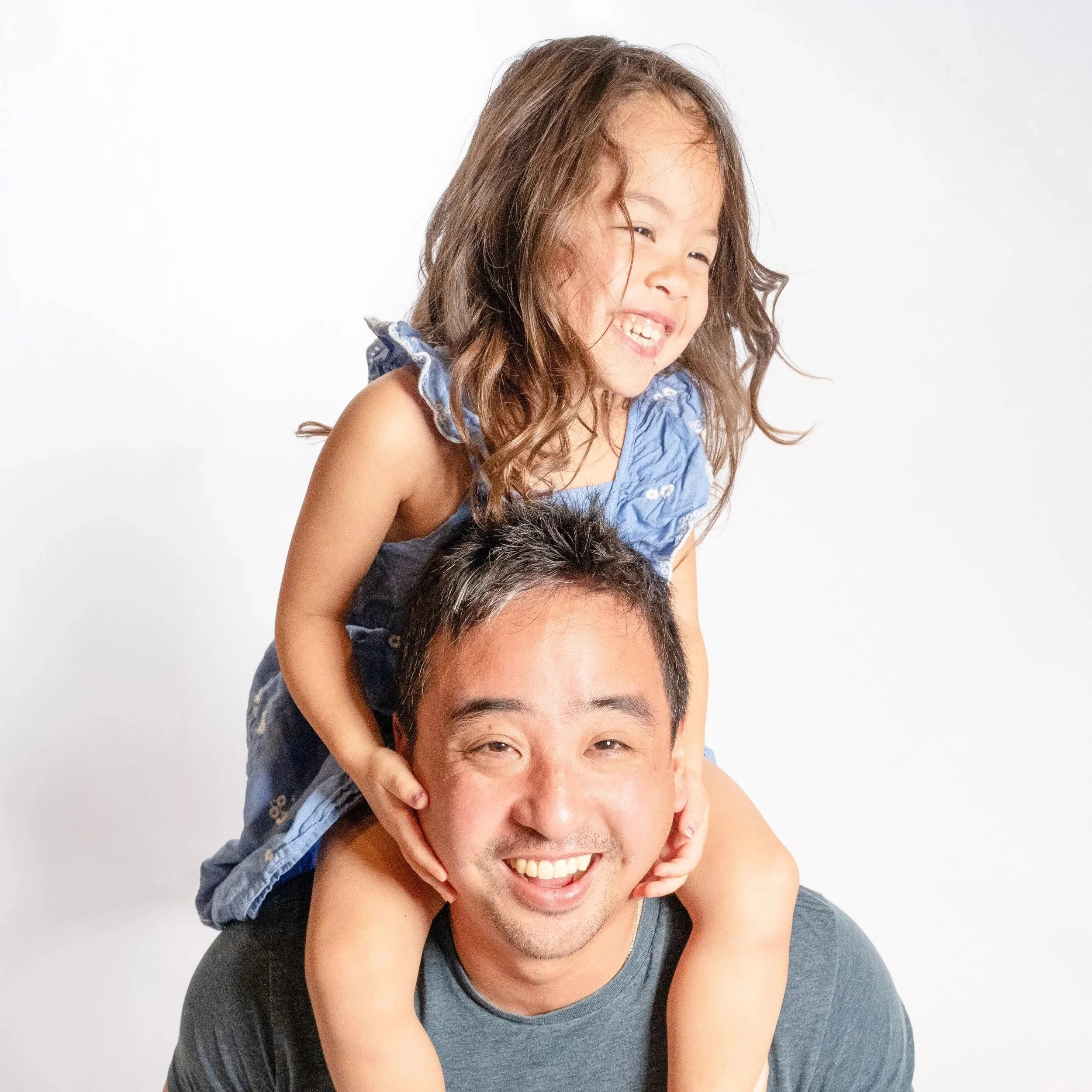 A young girl with long brown hair smiling and giving a thumbs-up while sitting on a man's her dad's shoulders, both against a white background located in Glenview, IL.