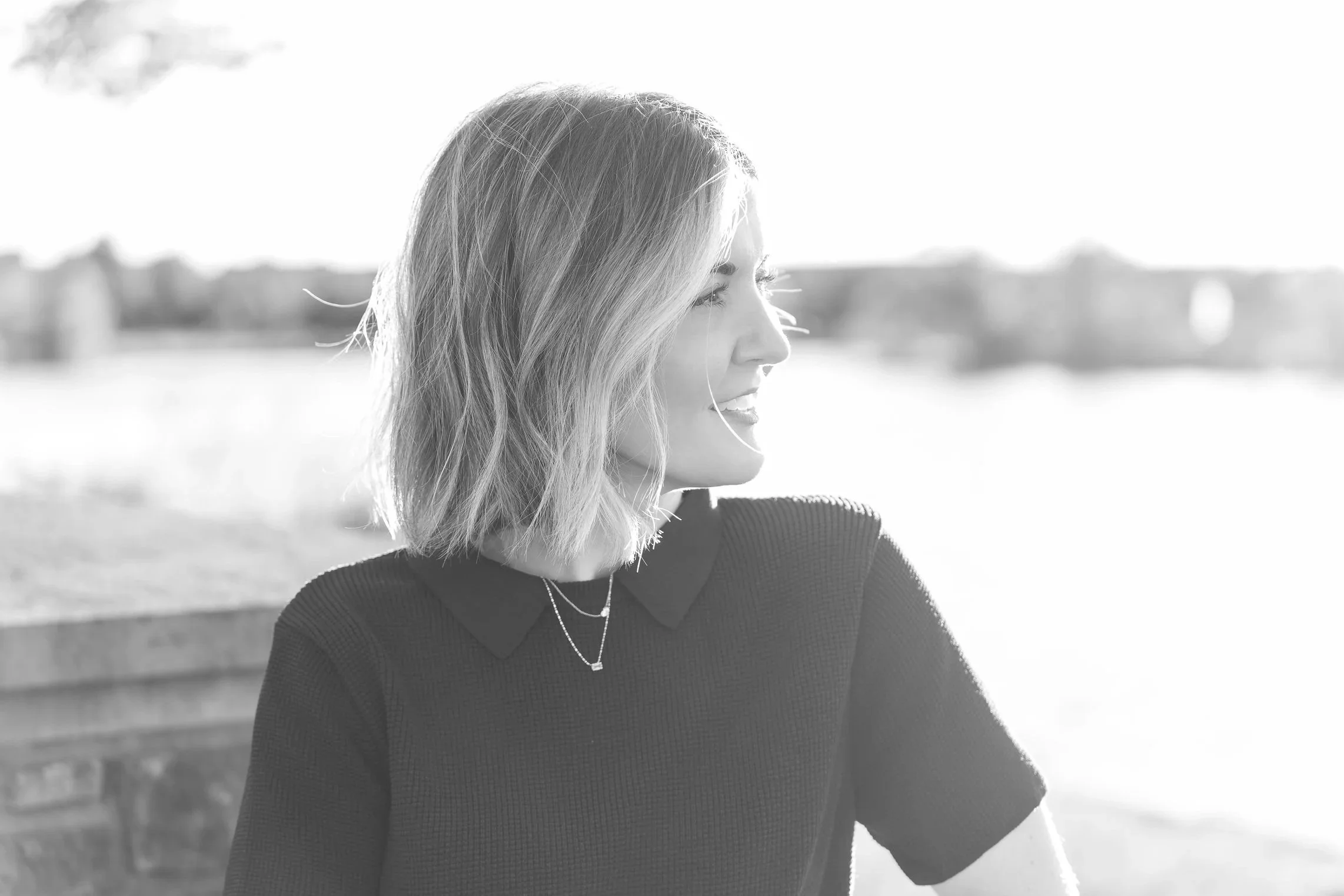 Black and white photo headshot of a smiling woman with shoulder-length wavy hair looking to the right, outdoors with blurred background located in the Glen in Glenview, IL.