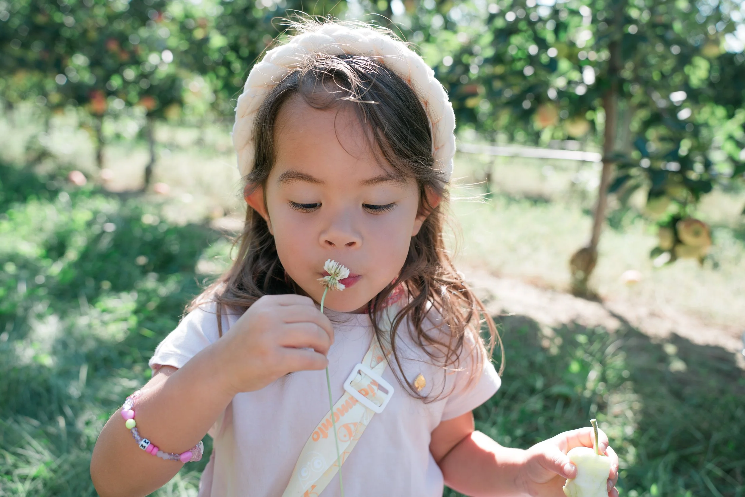 A young girl outdoors holding a white flower, wearing a pink shirt, with a forest background located on Edwards Apple Orchard.