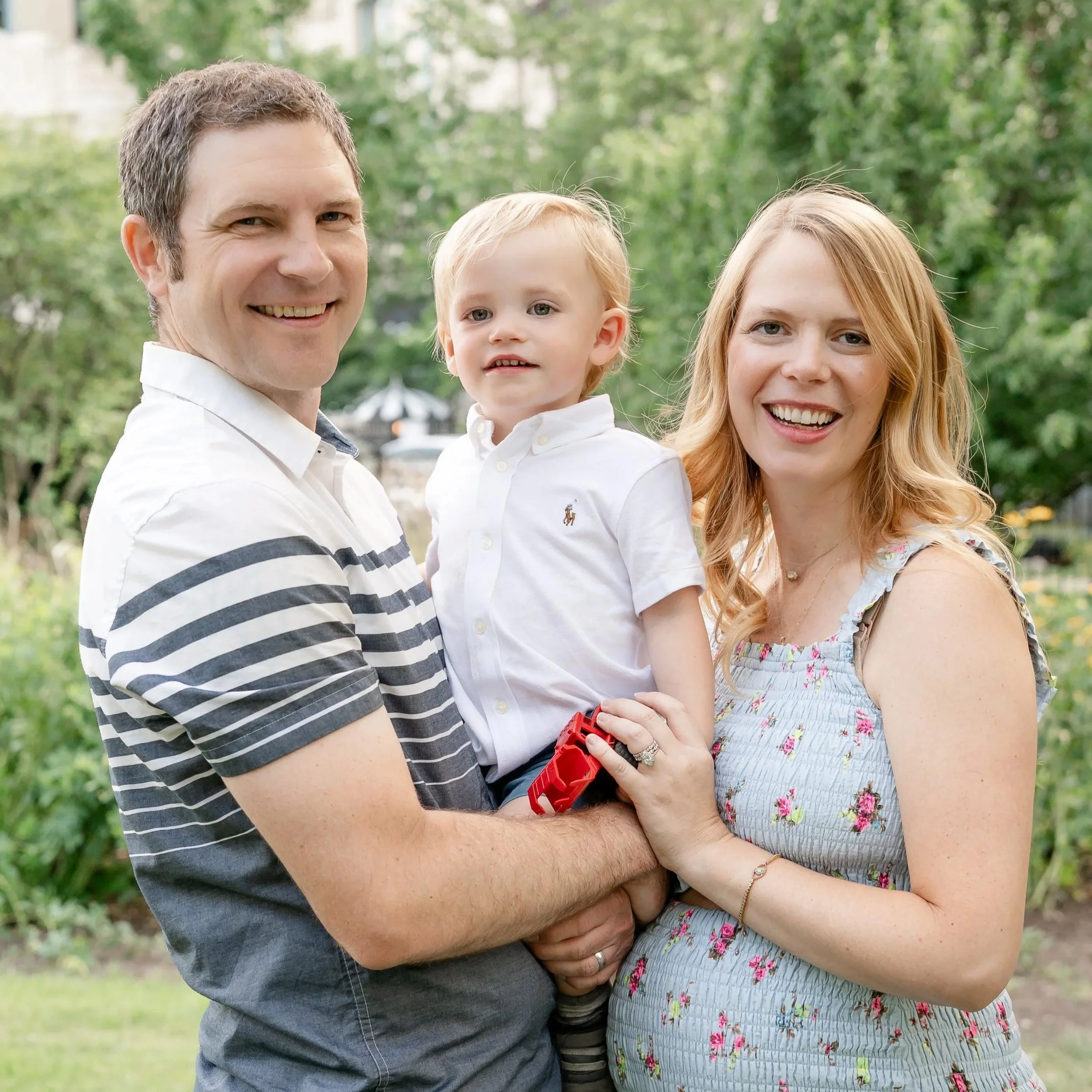 A man holding a young child and a woman standing beside them outdoors with trees in the background located in Lincoln Park Chicago