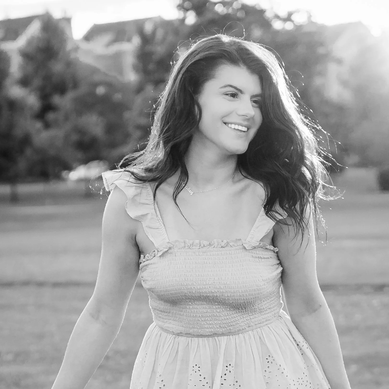 A young woman with long wavy hair smiling, wearing a light-colored dress with ruffled sleeves, outdoors with sunlight in the background located in The Glen in Glenview, IL.