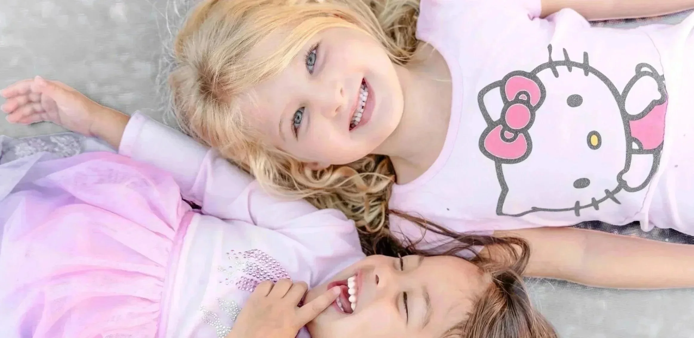 Three young girls smiling and lying on the ground together on a trampoline. They are dressed in pink and white clothes, with one girl wearing a shirt with Hello Kitty design. Located in Glenview, IL.
