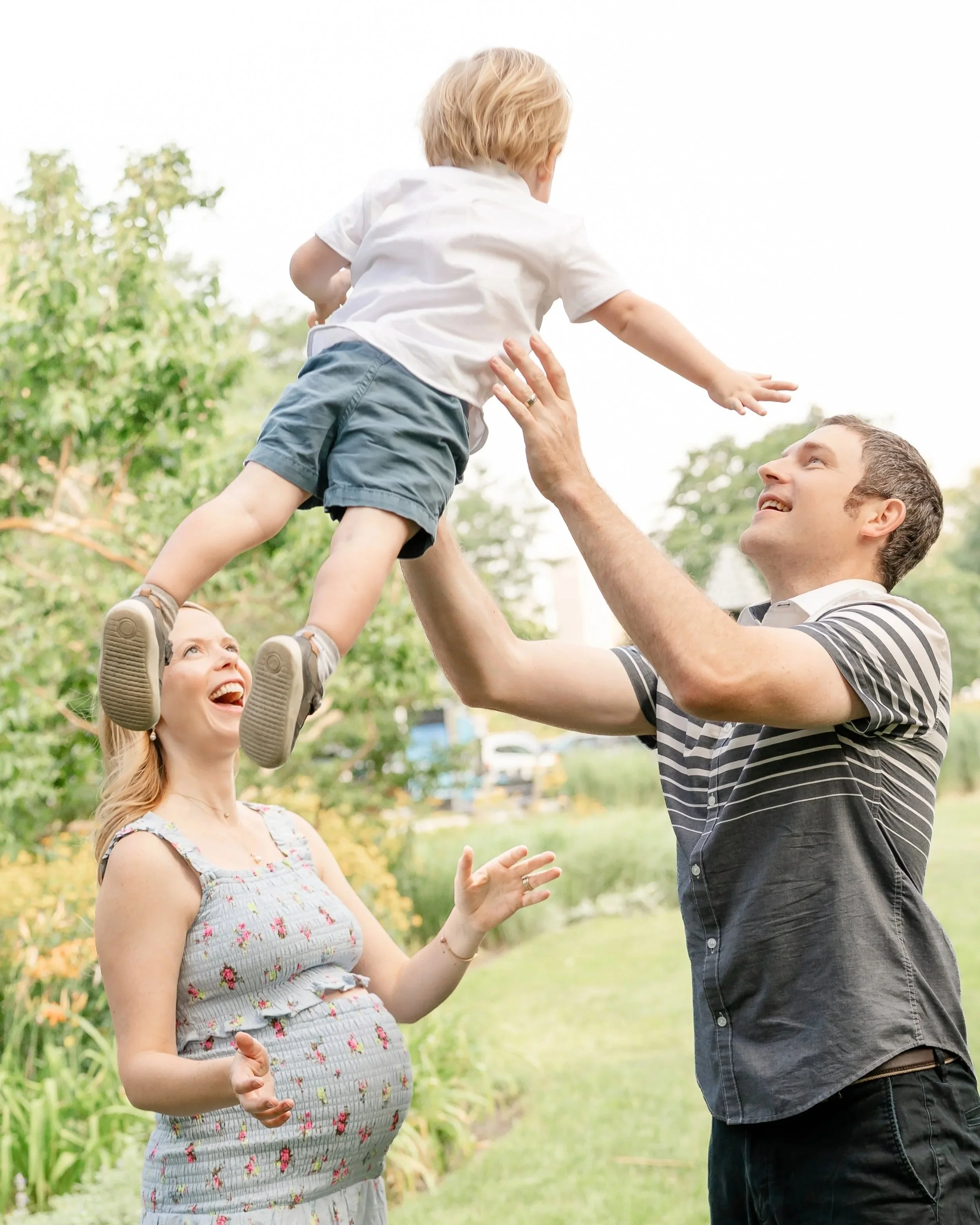 A man and a woman are outside playing with a young boy. The boy is being lifted into the air by the man, and the woman is smiling up at them, standing in a garden or park with trees and greenery in the background.