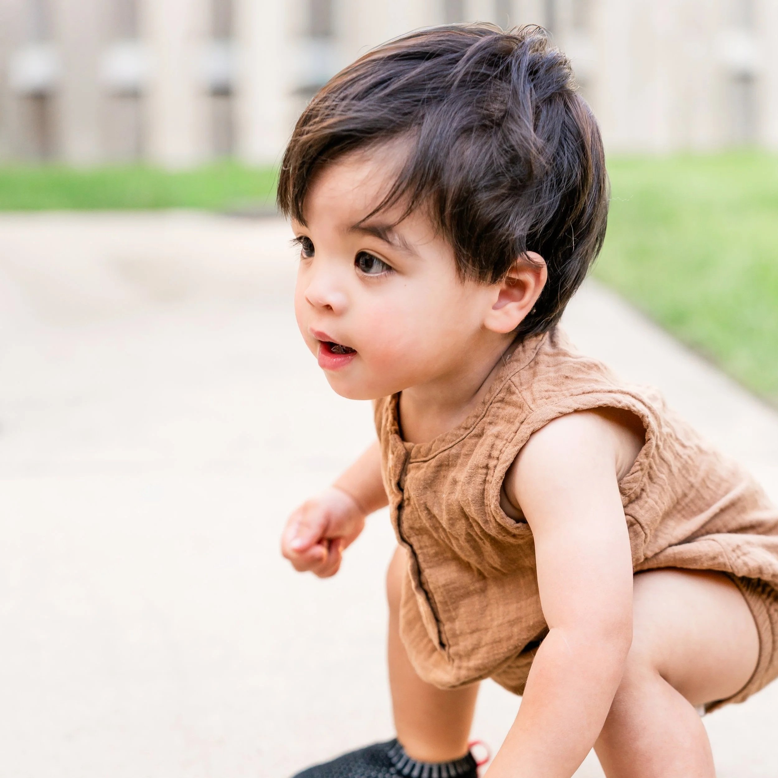 A young boy with dark hair, wearing a brown sleeveless shirt, sitting outdoors with a blurred background of grass and a building, looking to the left side of the image located in Glenview, IL.