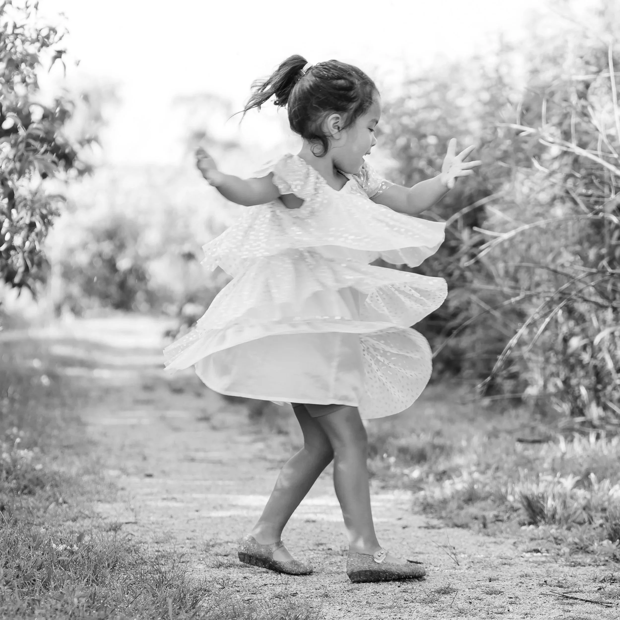 A young girl playing on a large piece of fabric and dress outdoors, standing on one leg with arms extended, surrounded by grass and blurred natural background in The Glen in Glenview, IL.
