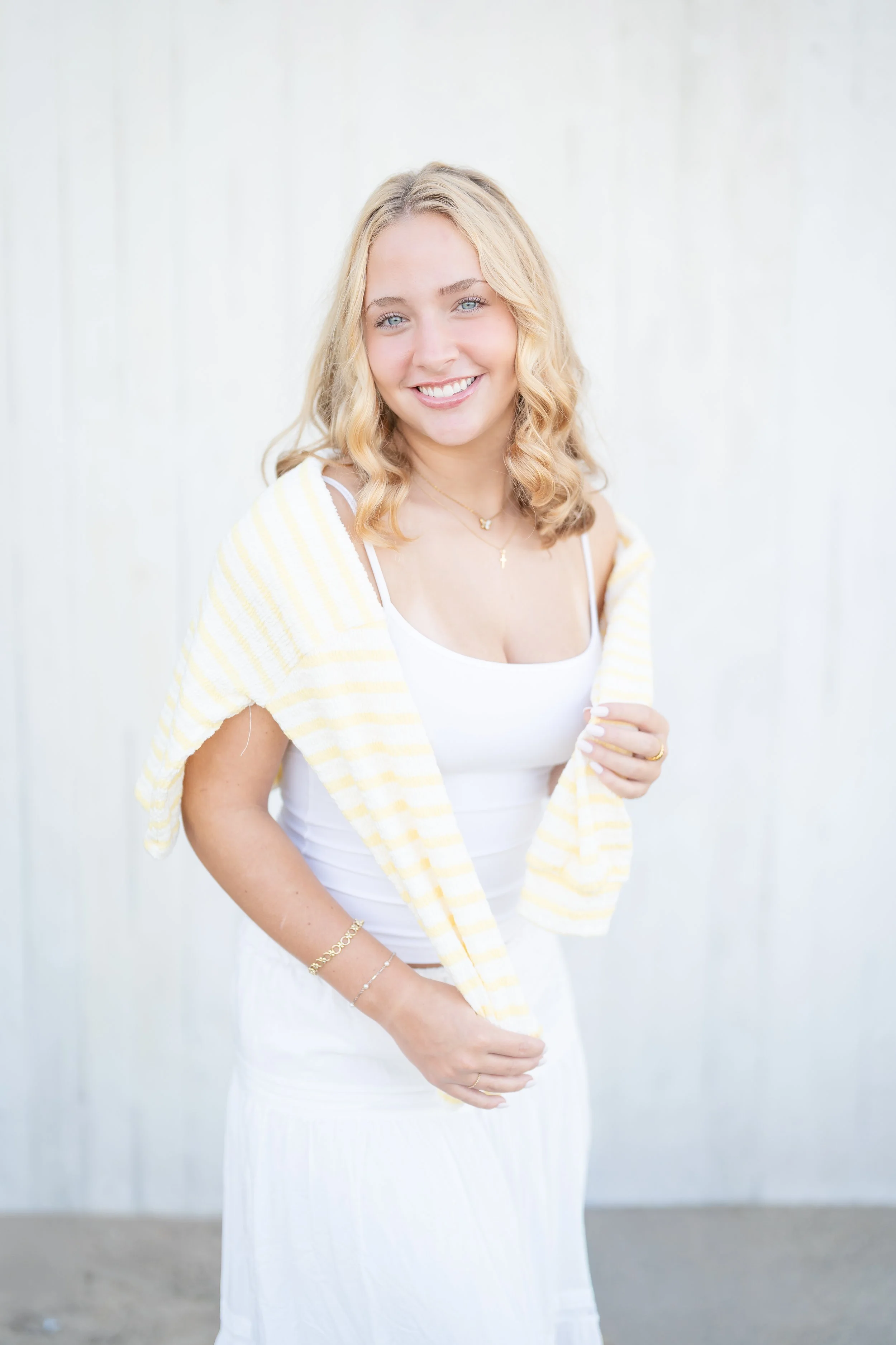 A young woman with blonde hair and blue eyes smiling while wearing a white dress and a yellow striped cardigan, standing indoors with a white wall background, located in Wilmette at Gillson Beach.