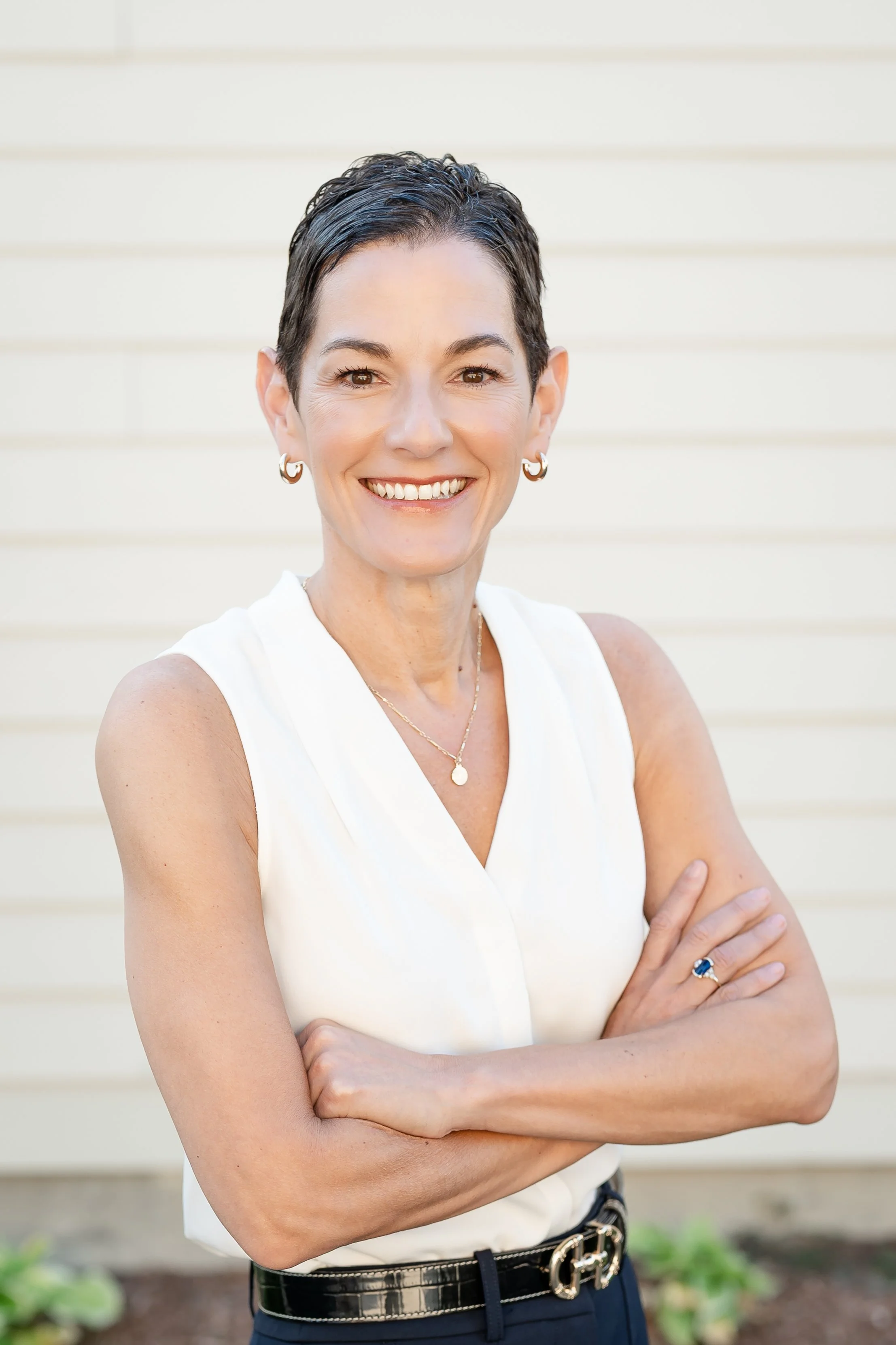 A woman with short dark hair, smiling, wearing a white sleeveless top, gold hoop earrings, a gold necklace, a ring with a blue stone, and a designer belt, standing outdoors against a light-colored horizontal siding background in Glenview IL.