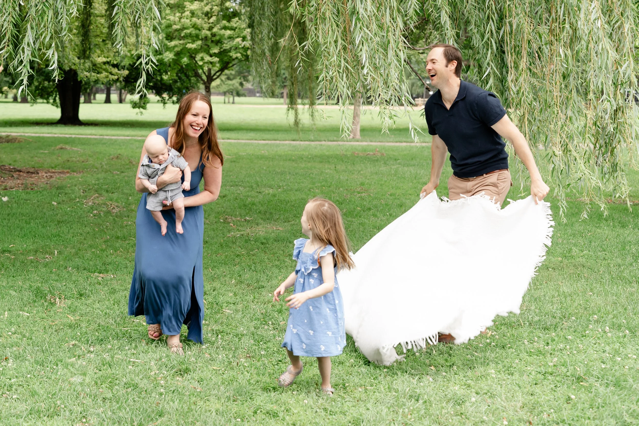 A family enjoying outdoor activities in a park with green grass and trees. A woman is holding a baby, two young girls are walking, and a man is smiling and holding a large white fabric or blanket located in Glenview IL.