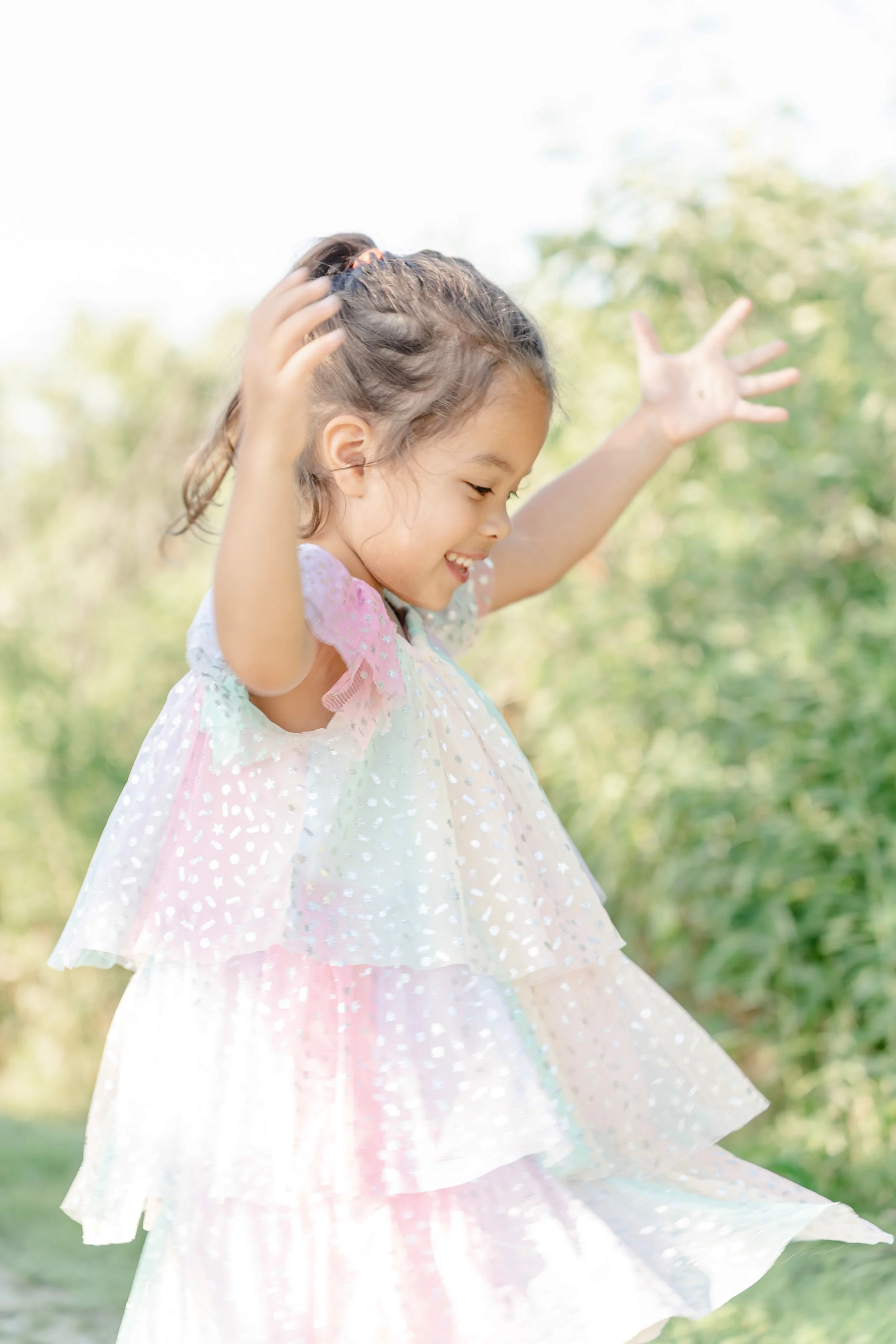 Young girl outdoors wearing a flowing dress, smiling, with arms raised and hands open, surrounded by greenery located in the Glen in Glenview, IL.