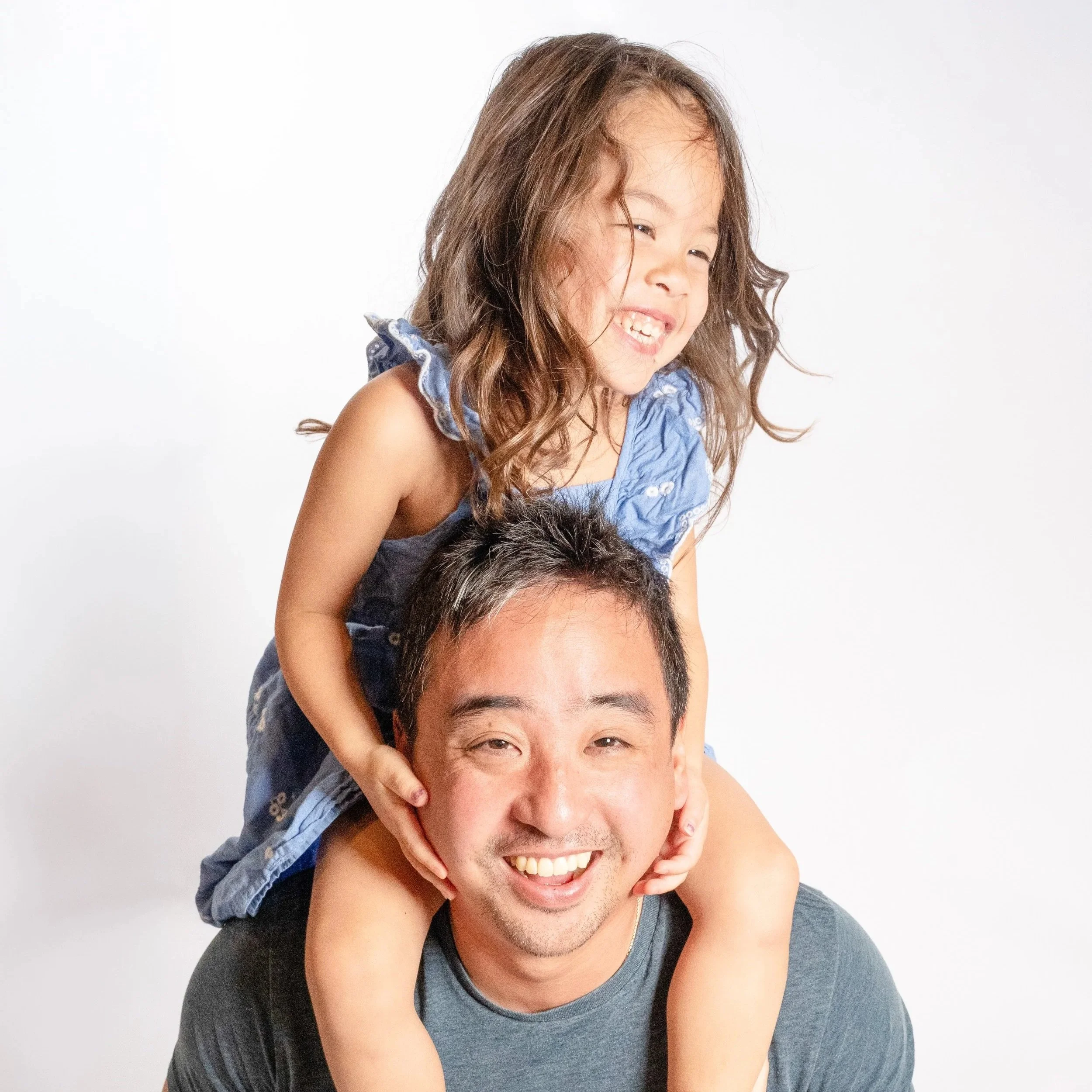 A young girl with long brown hair smiling and sitting on a man's shoulders, who is also smiling, both against a plain white background located in Glenview IL.