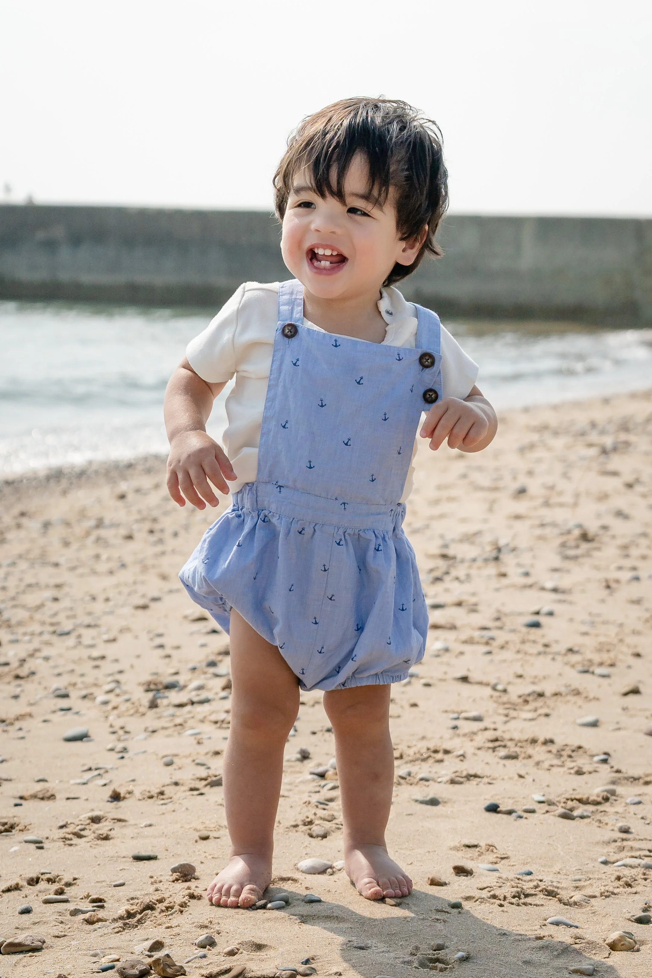 A young child with dark hair and wearing a white shirt and blue overalls with small anchors prints, standing barefoot on a sandy beach with rocks, smiling and running located in Glencoe IL
