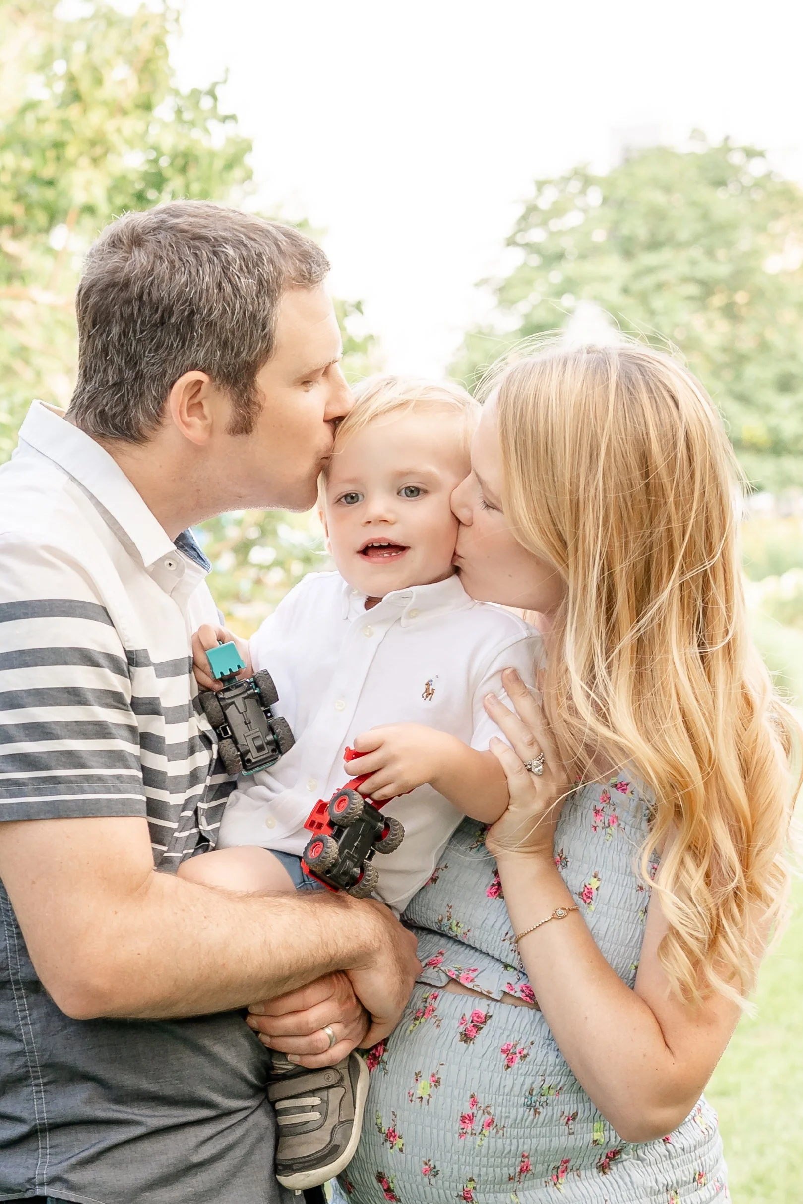 A family outdoors, with a man and woman kissing their child on the cheeks, holding toy cars, in a park with trees in the background located in Winnetka, IL.