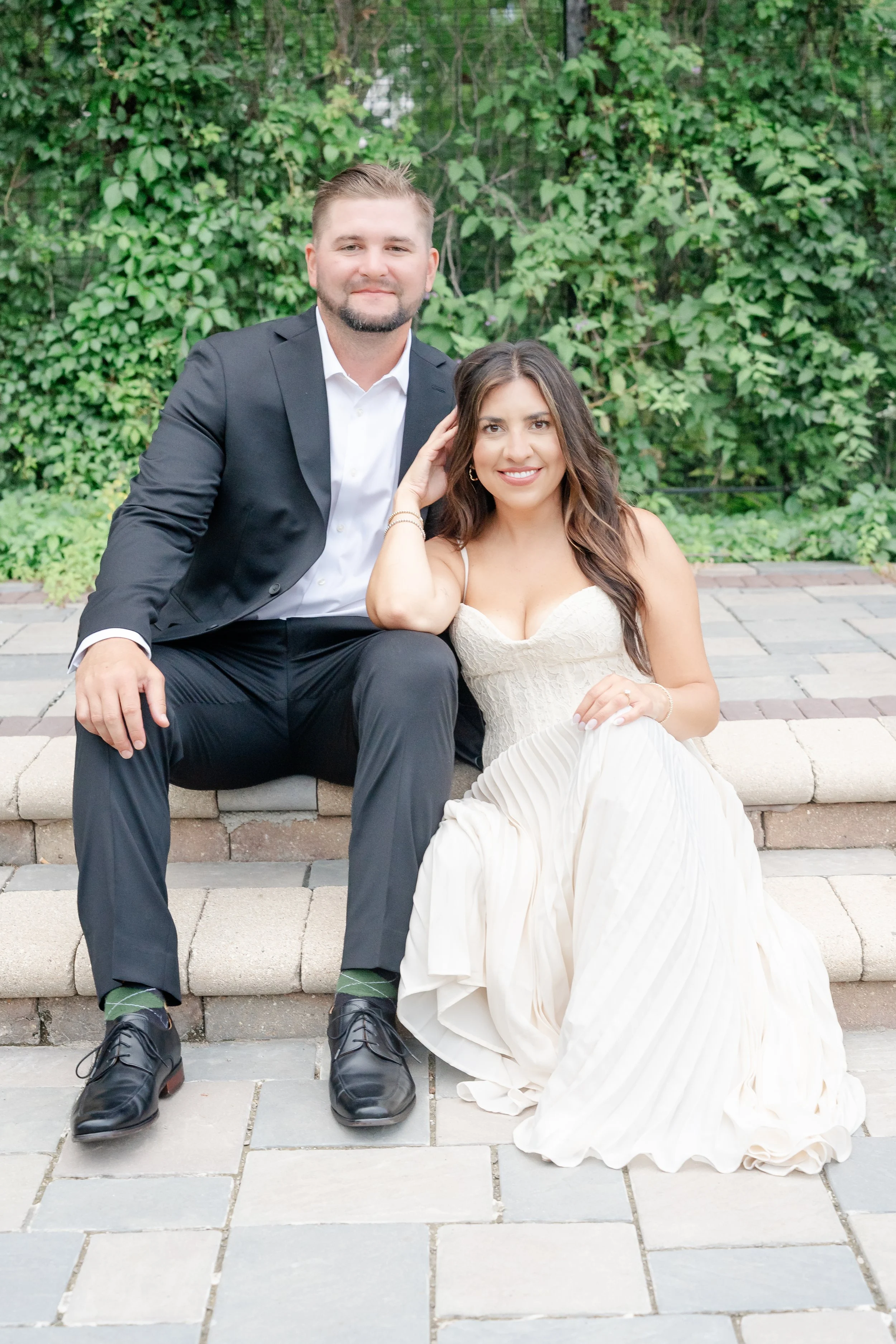 A man and woman sitting on a stone bench outdoors, with green foliage behind them. The man wears a black suit and white shirt, and the woman wears a cream-colored dress in Glenview IL