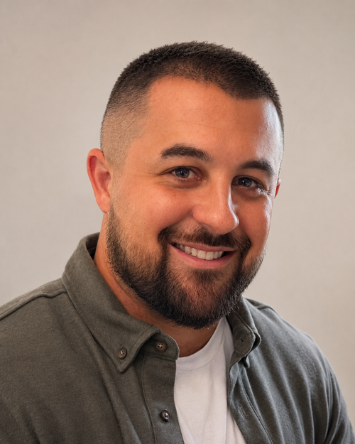 Portrait of a smiling man with short dark hair, beard, wearing a gray button-up shirt over a white t-shirt, against a plain neutral background.