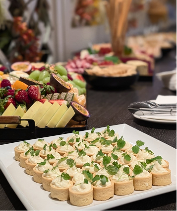 Tray of small appetizers with creamy topping and green herbs in the foreground, assorted fruits, cheeses, and meats on platters in the background.