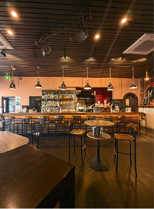 An empty bar or restaurant with a wooden bar counter, liquor bottles on shelves, multiple hanging pendant lights, and several small round tables with chairs, in a cozy setting.