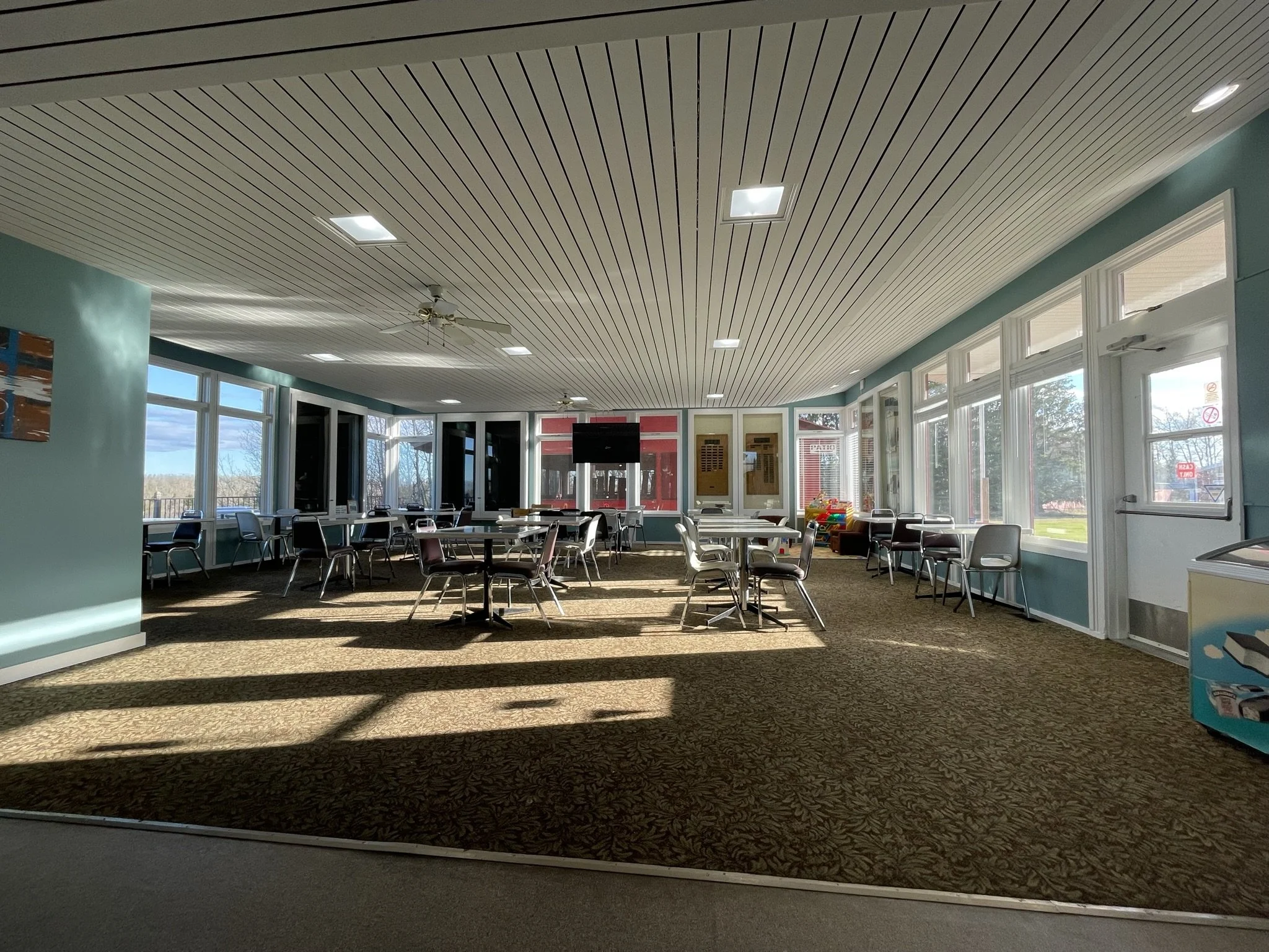 Indoor space with large windows, ceiling fans, tables, and chairs, with sunlight casting shadows on a patterned carpet.