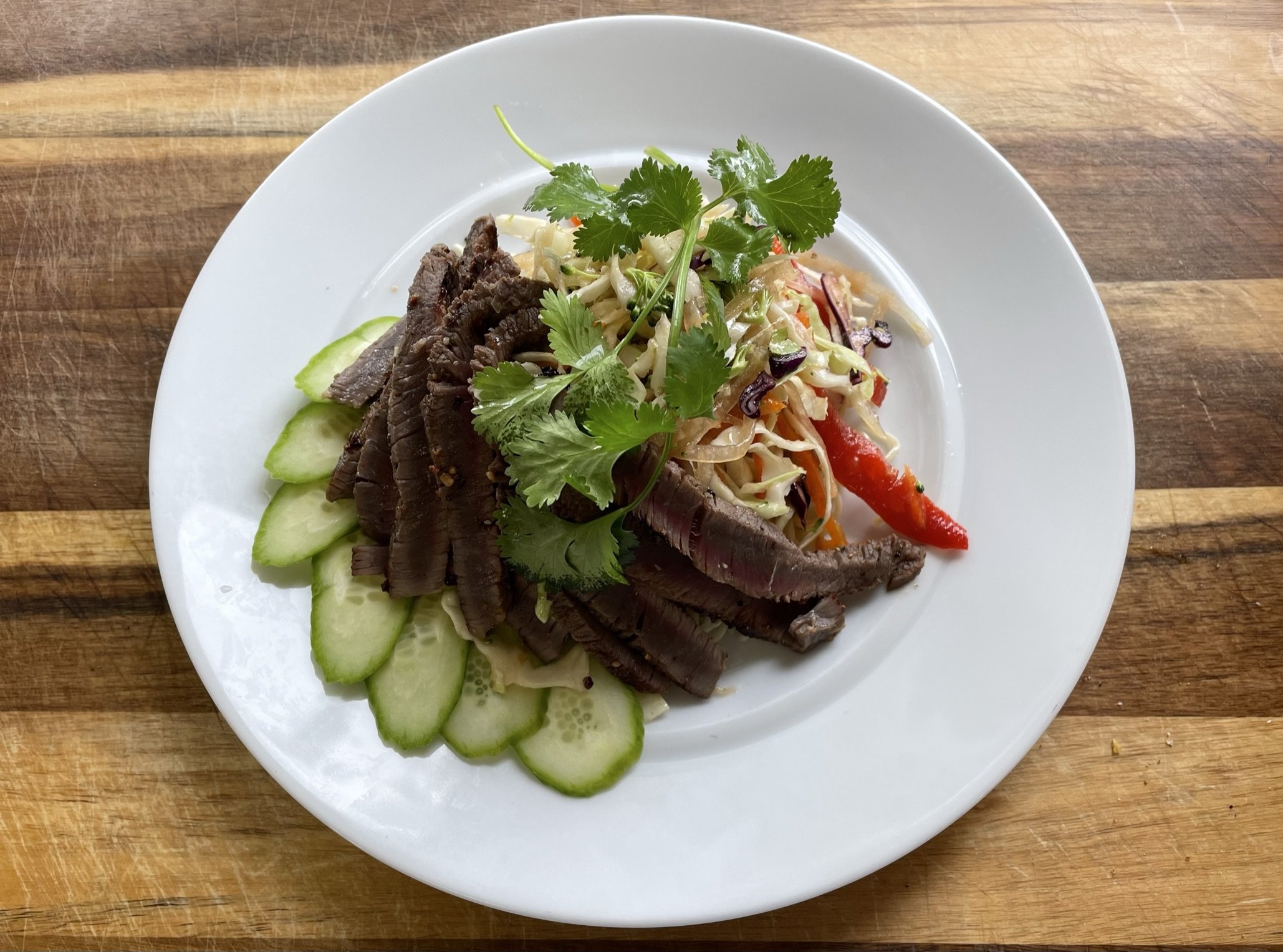 A white plate of sliced beef, shredded cabbage salad with carrots, peppers, and cilantro, and sliced cucumber placed on a wooden table.