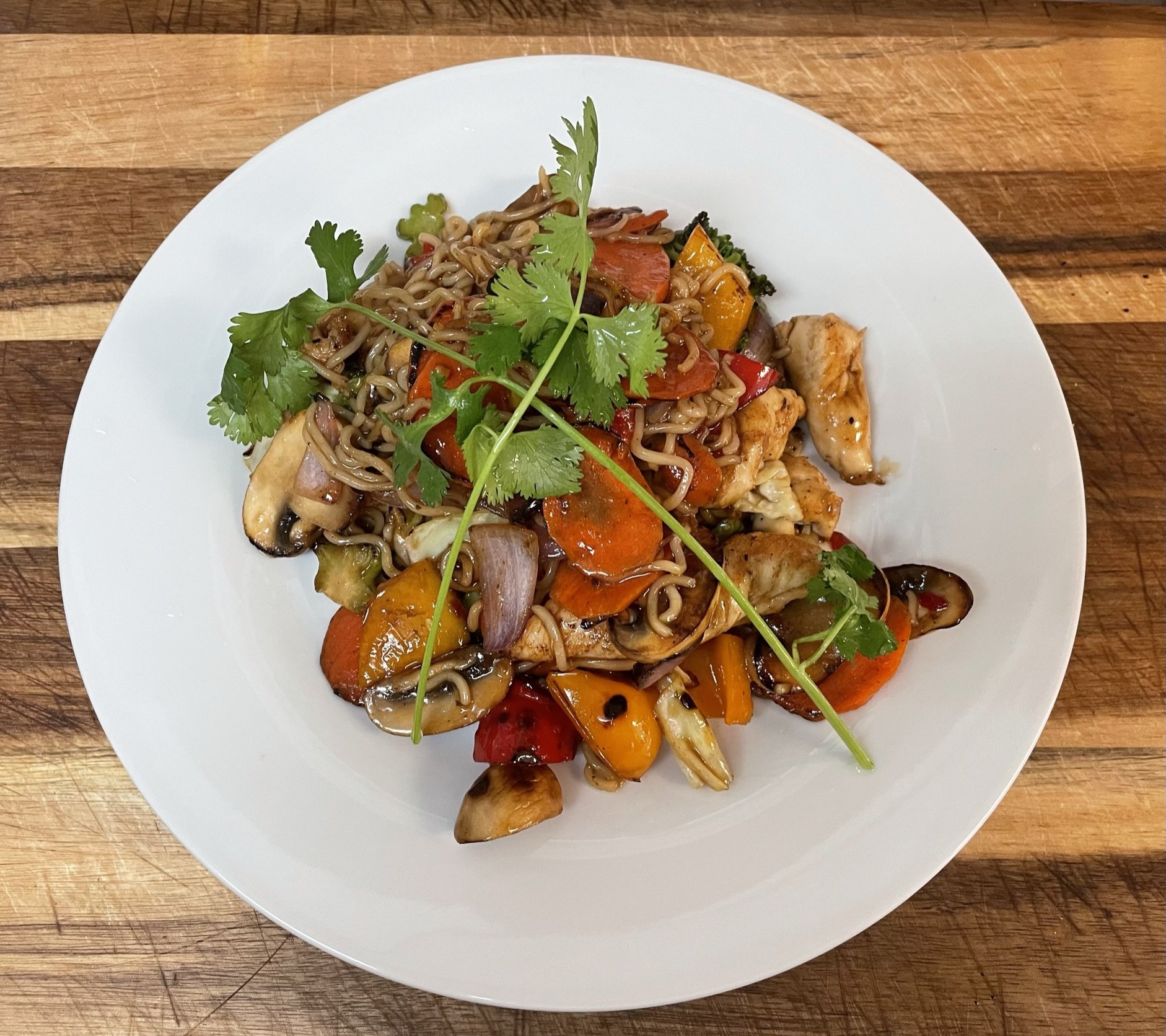 A white round plate with stir-fried vegetables and noodles garnished with cilantro, placed on a wooden surface.