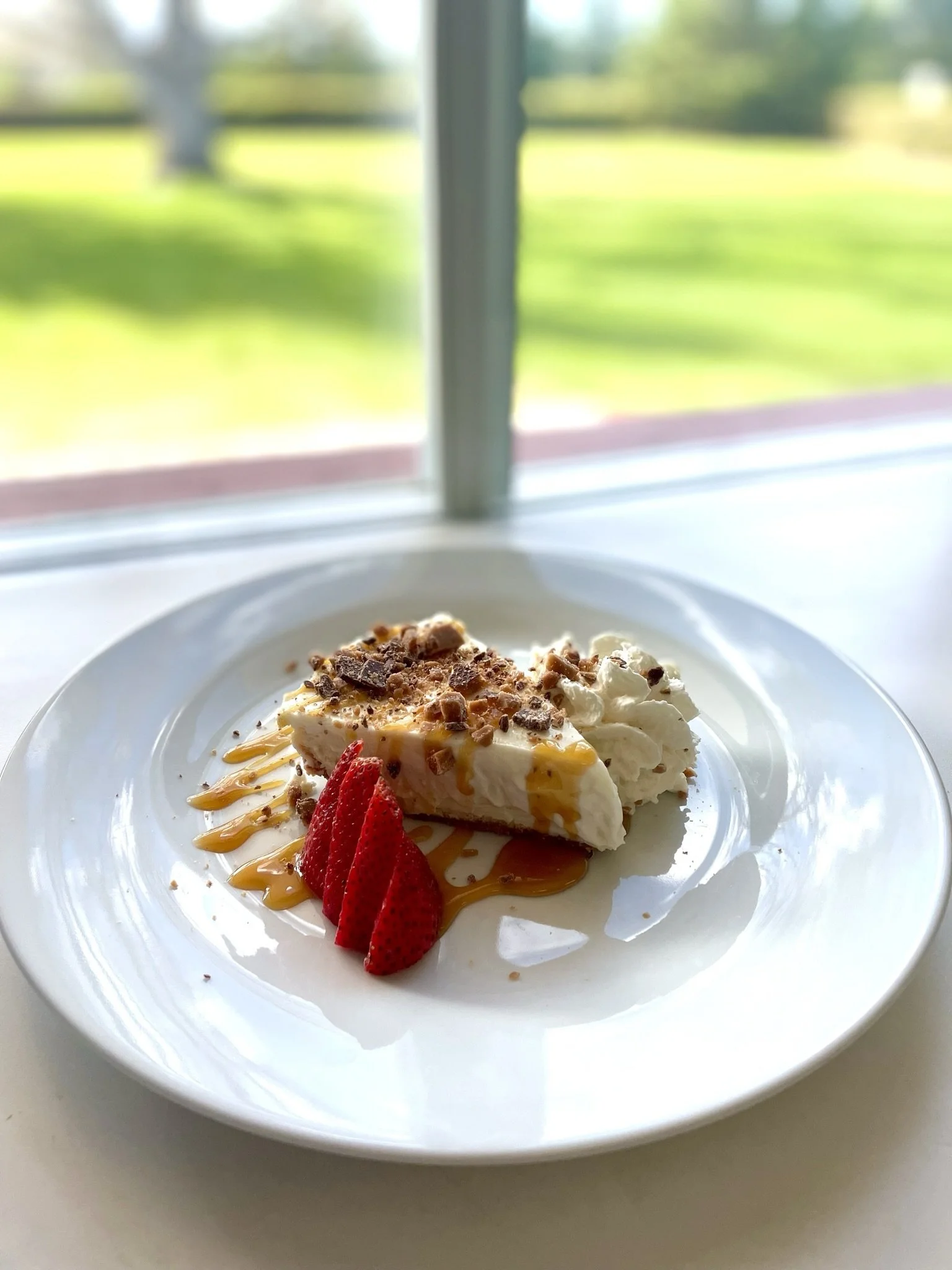 Slice of cheesecake topped with whipped cream, caramel drizzle, chopped chocolate, and fresh strawberries in a white bowl, with a window and outdoor scenery in the background.