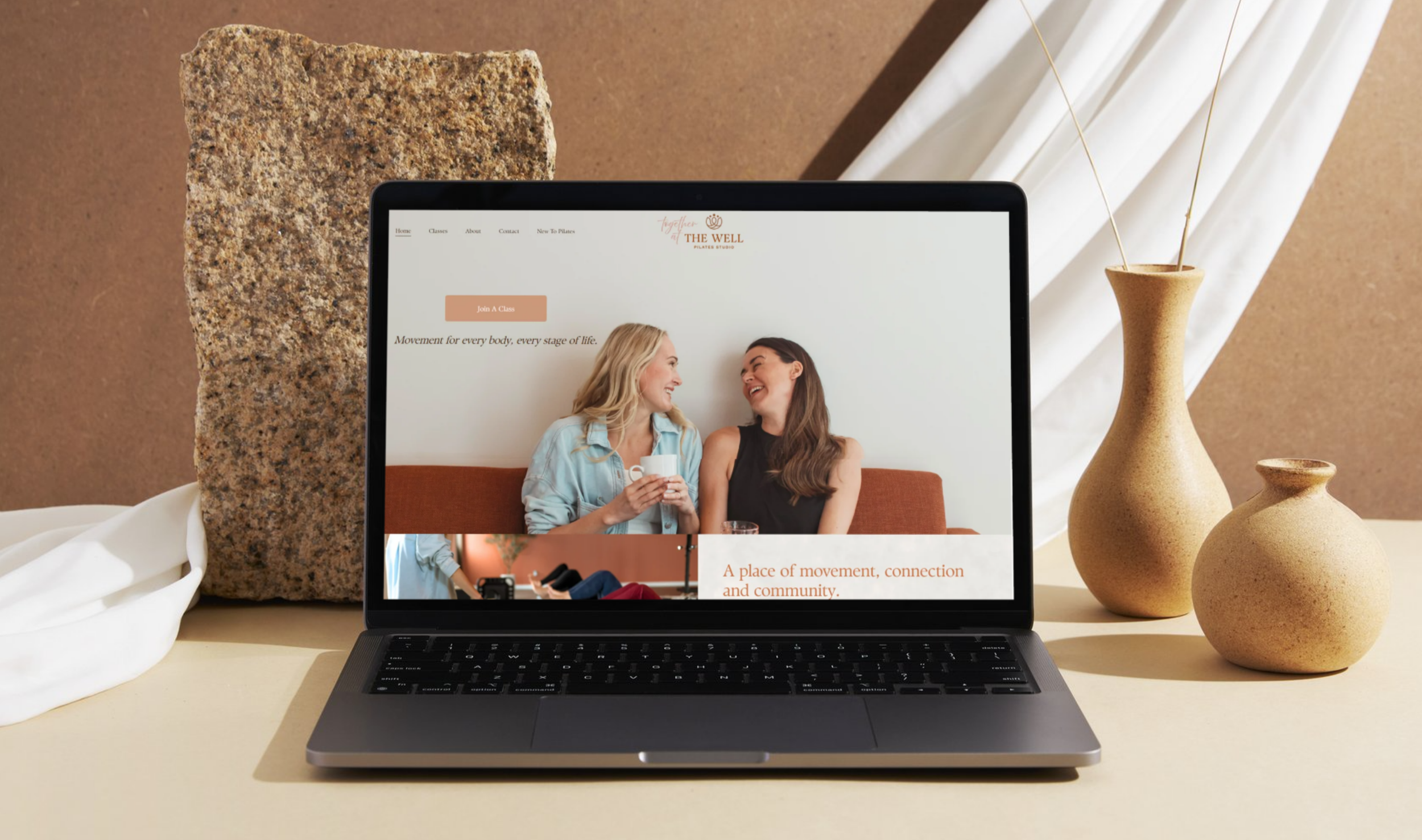 Laptop displaying a website for The Well Pilates Studio, featuring a photo of two women laughing and conversing, with pottery and textured brown and white walls in the background.