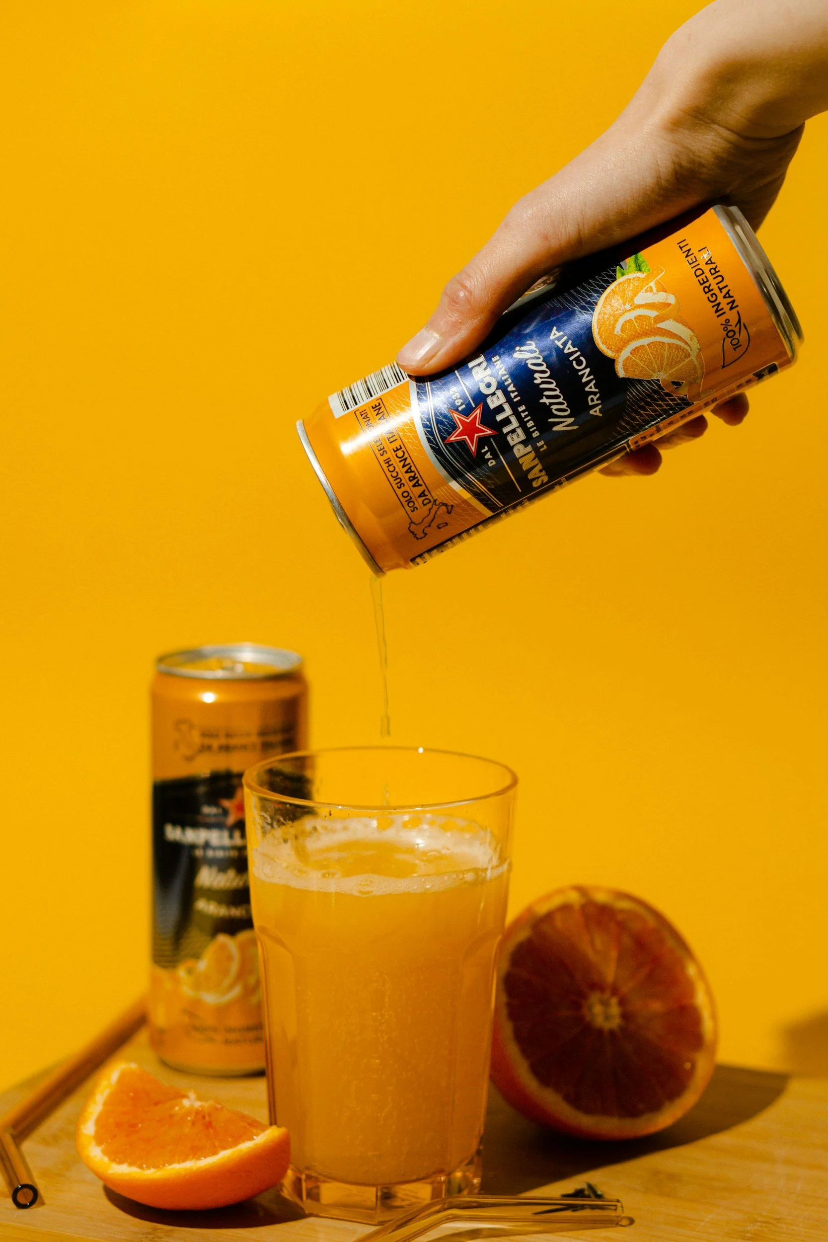 A hand holding a can of sparkling orange flavored beverage above a glass filled with the drink. Whole and sliced oranges are on the table, set against a yellow background.