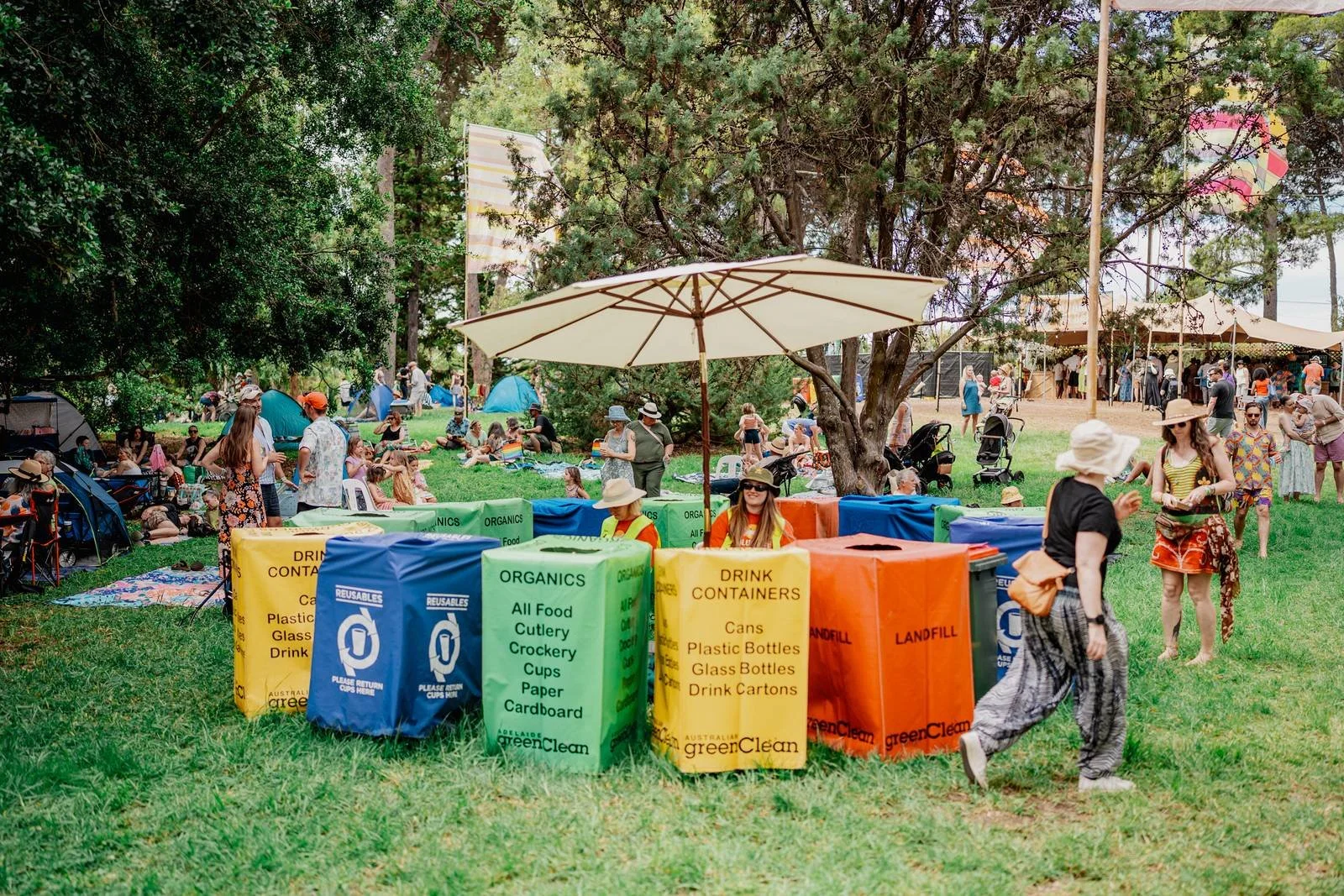A community event in a park with numerous people, trees, and a recycling station with colored bins labeled for recyclables and landfill.