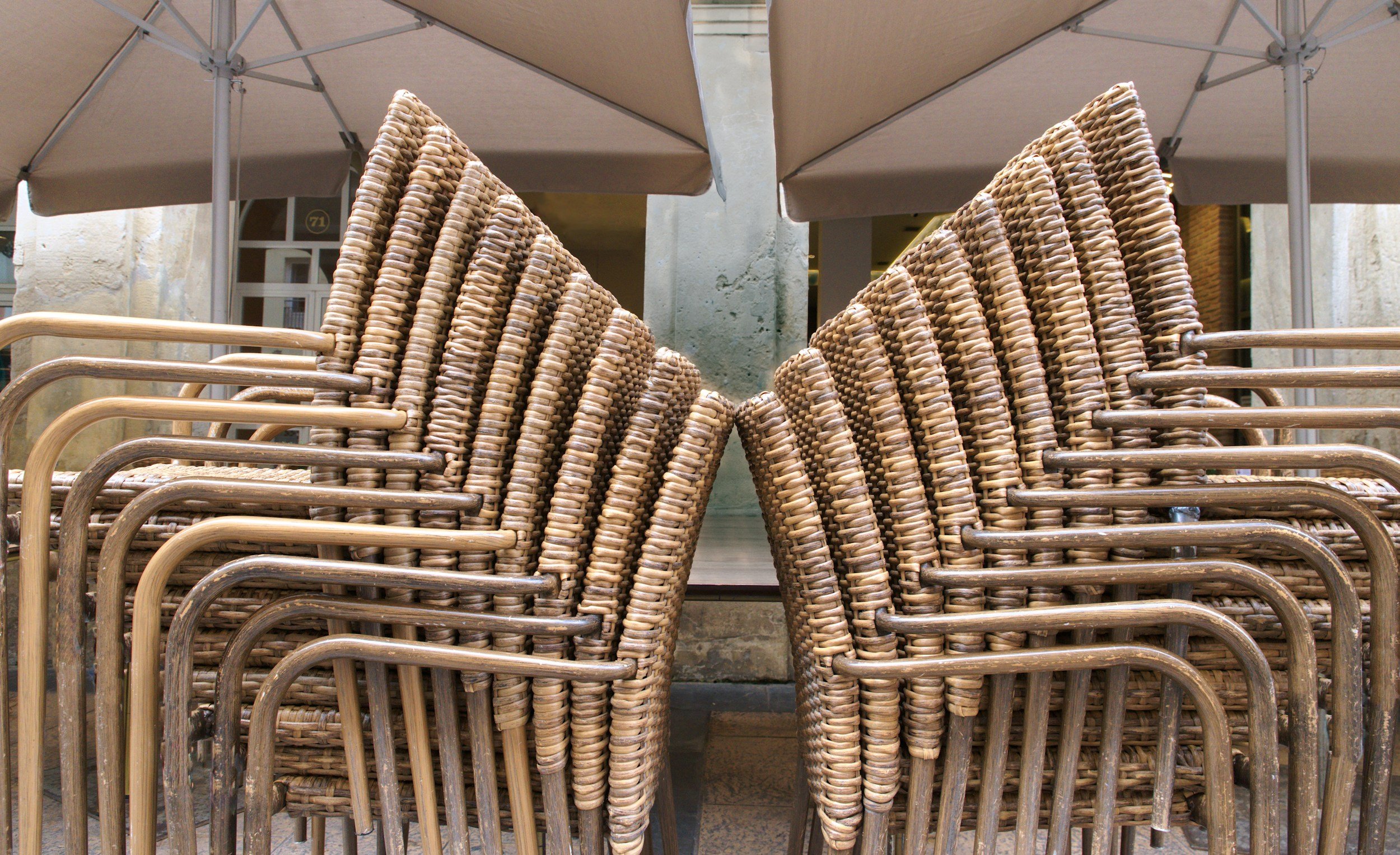 A stack of outdoor woven rattan chairs with metal frames underneath large beige patio umbrellas.