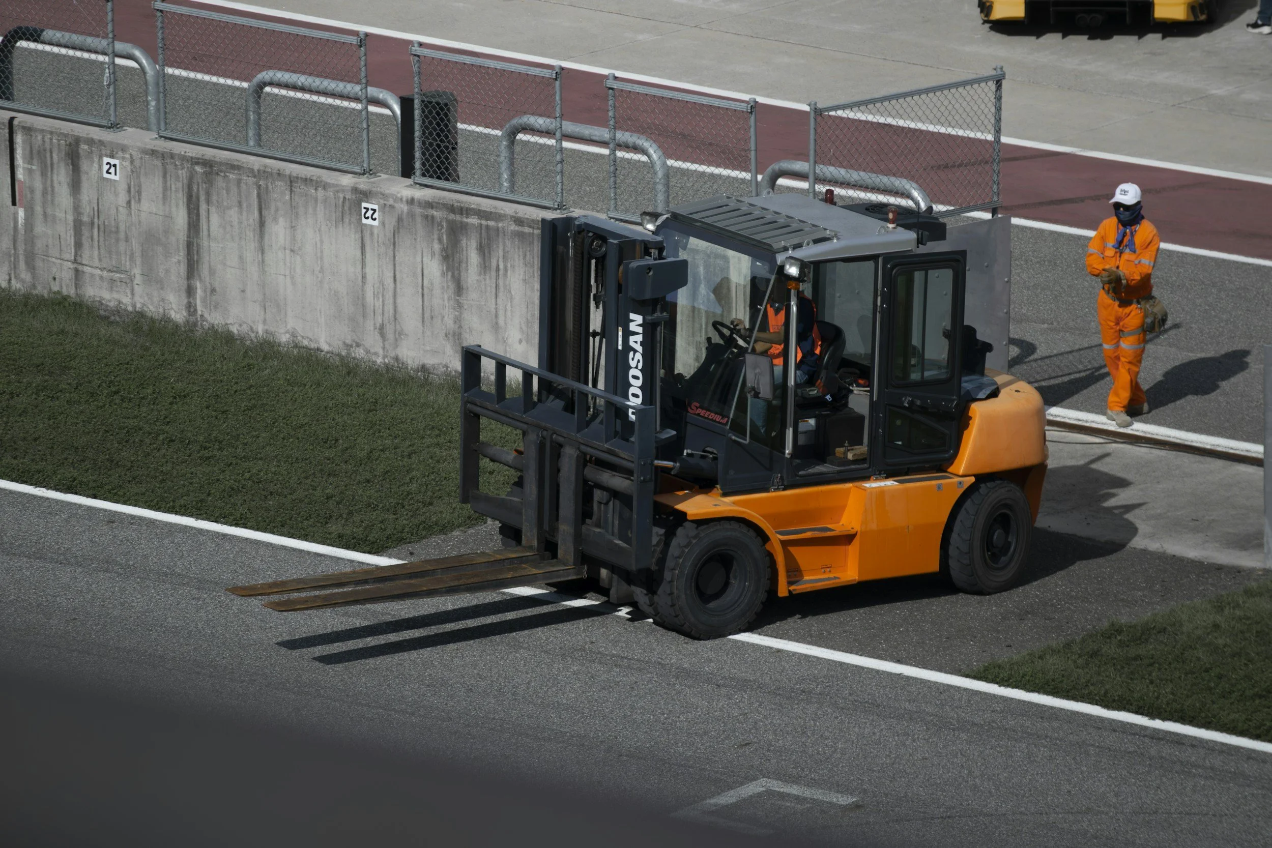 A forklift operator in an orange jumpsuit and a person in an orange jacket and white cap and face mask are working near a concrete barrier at a sports stadium or track field. The forklift is lifting a section of the barrier's fencing.