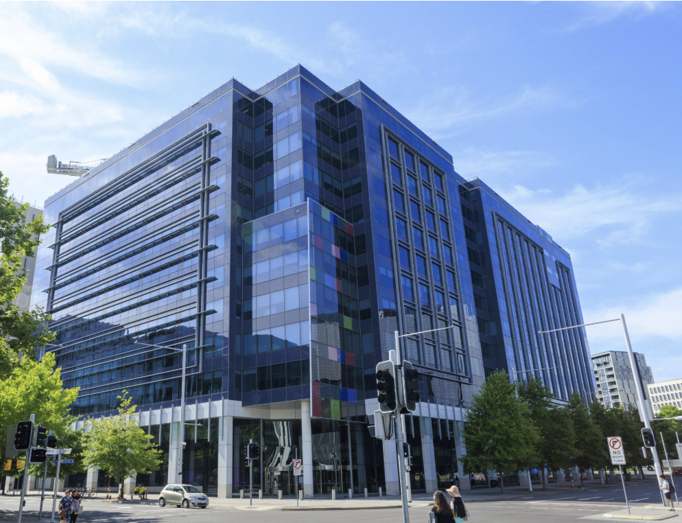 Modern glass office building with blue-tinted windows, surrounded by greenery and city street with pedestrians and traffic lights.
