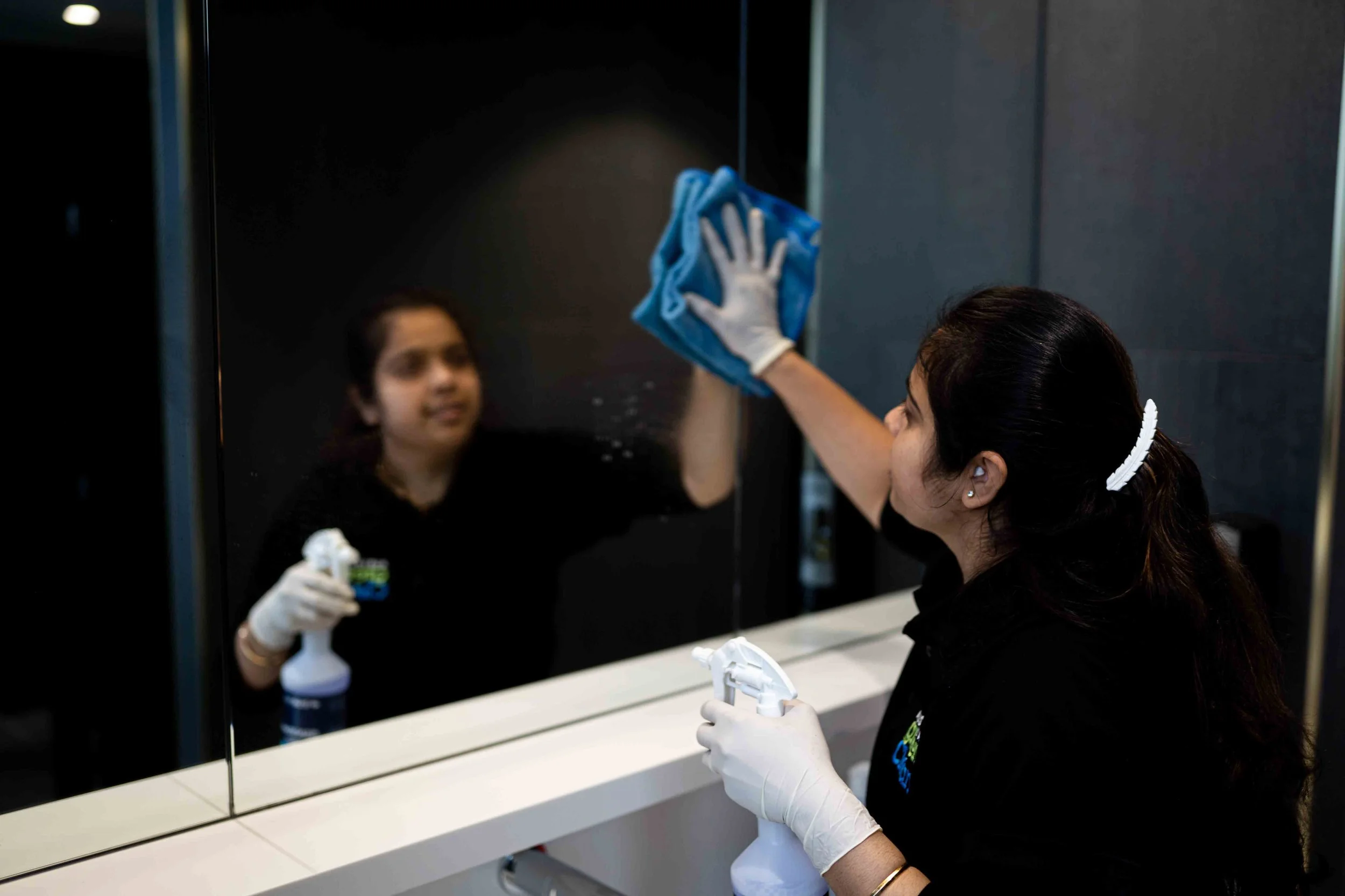 A woman wearing gloves is cleaning a mirror in a bathroom or similar setting. She is using a spray bottle and a blue cloth, and her reflection is visible in the mirror. The countertop below is white, with cleaning supplies nearby.