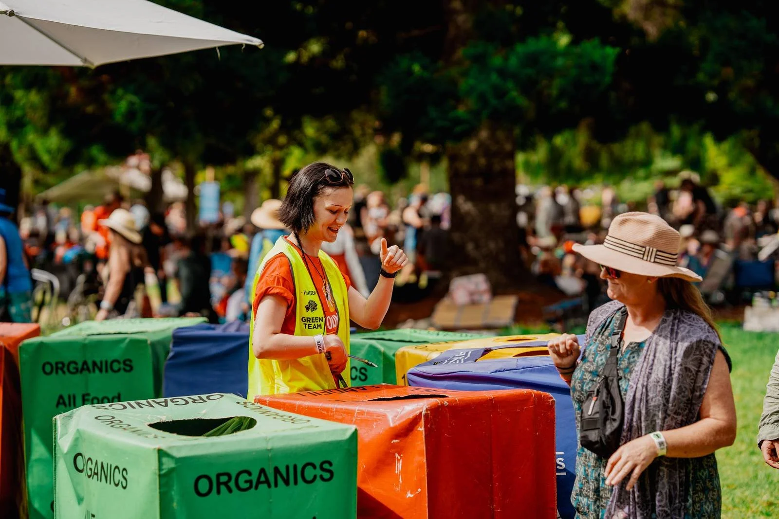 A woman with short black hair, wearing a red shirt and a yellow volunteer vest, is smiling and gesturing with her thumb up while talking to another woman in a wide-brimmed hat at an outdoor event. There are green, blue, yellow, and orange bins labeled 'ORGANICS All Food' around them, and a crowd of people in the background under trees.